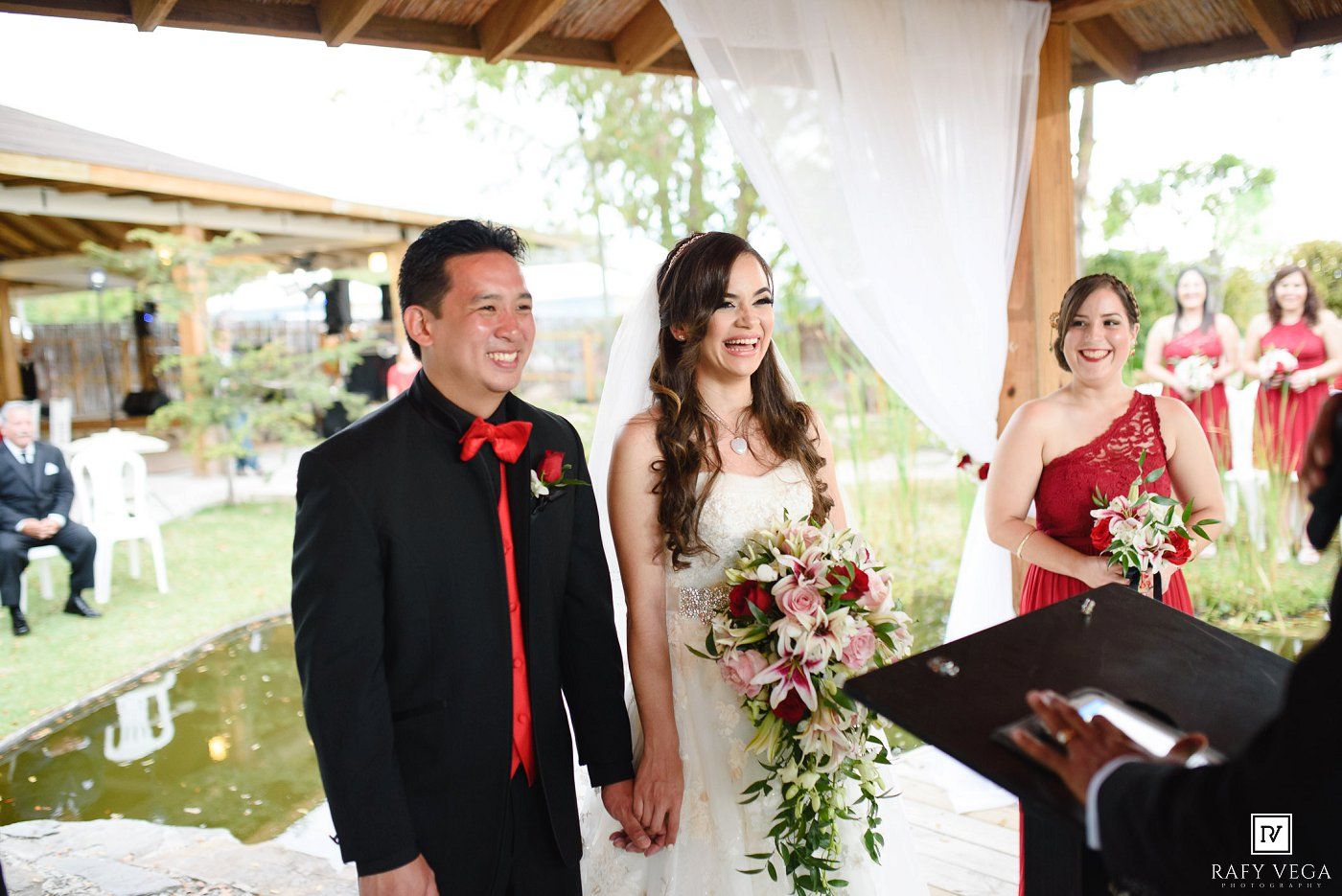 Bride and groom smiling during outdoor wedding ceremony, bride holding red and white bouquet, officiant in red dress nearby.