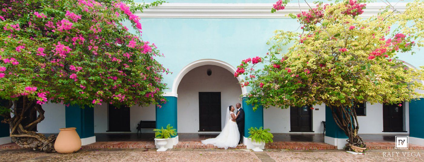 Couple shares romantic wedding moment under turquoise archway surrounded by vibrant pink bougainvillea trees.