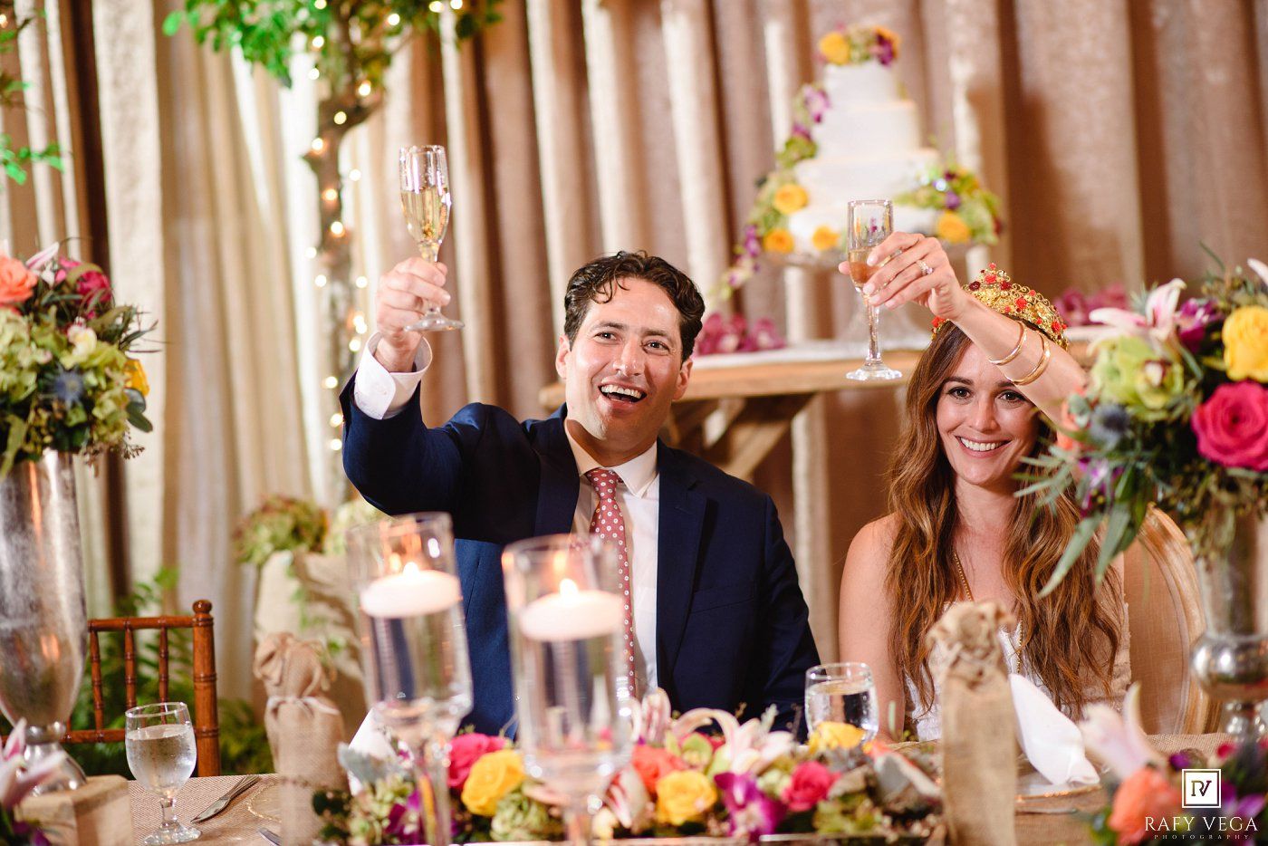 Bride and groom raising champagne glasses in a toast at their elegant wedding reception with colorful floral arrangements.