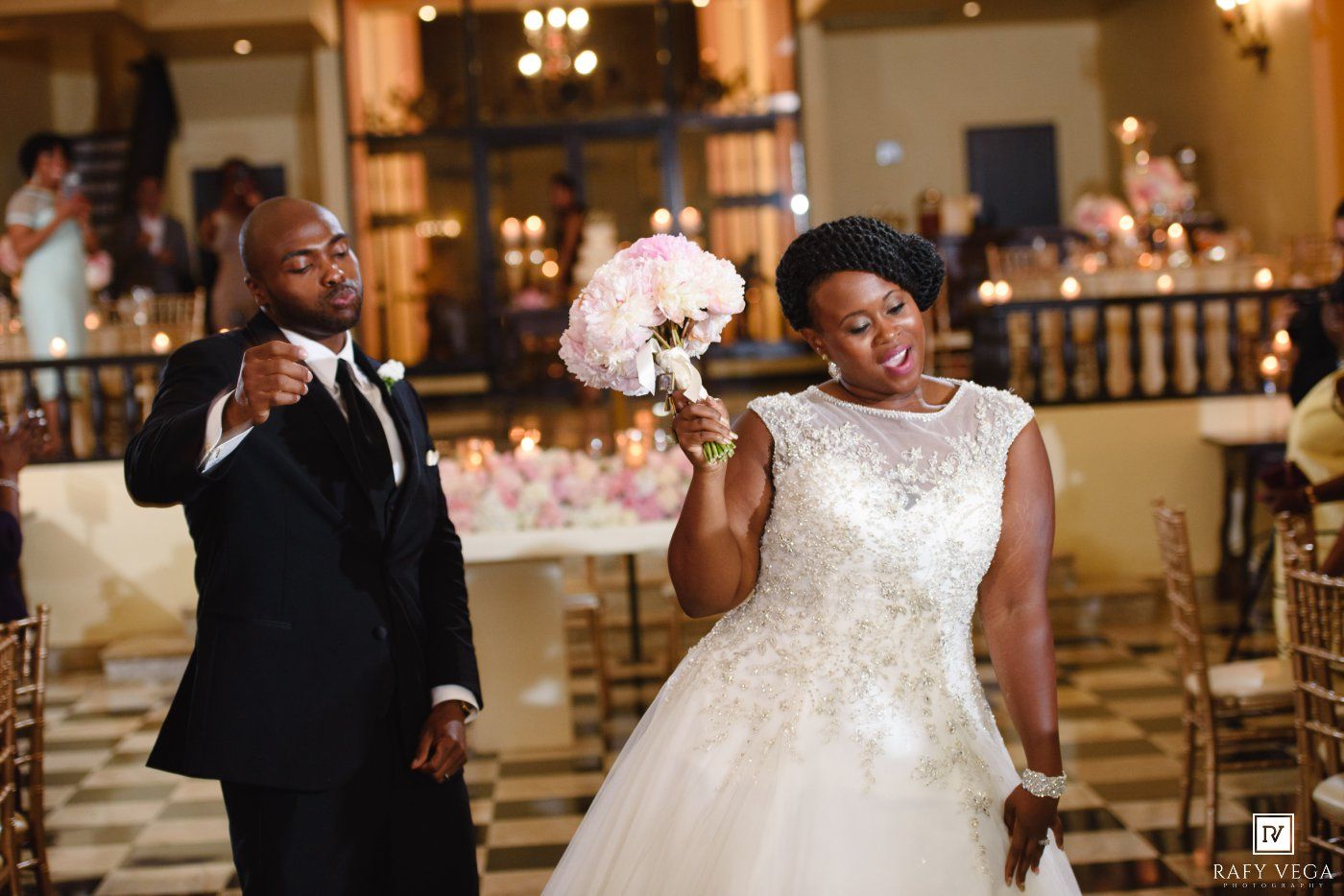 Bride in white beaded gown holds pink bouquet aloft as groom in tuxedo gestures during elegant wedding reception.