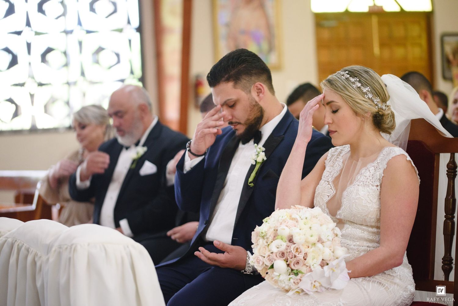 Bride and groom sit together in church pews during wedding ceremony, bride holding white floral bouquet.