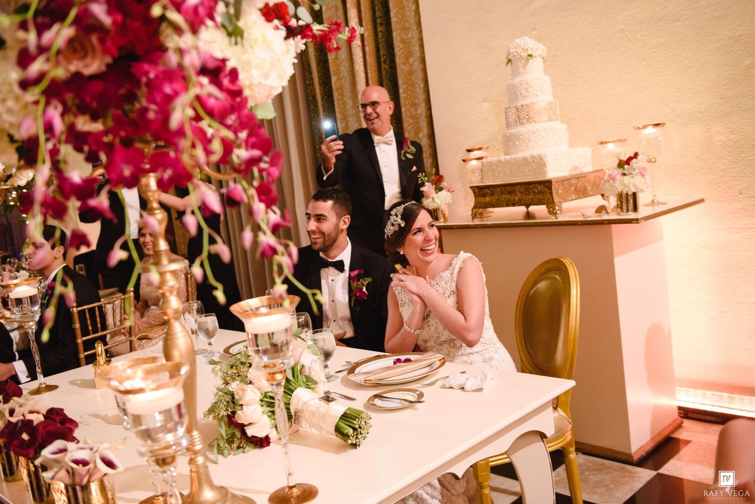 Bride and groom seated at elegant wedding reception table with gold accents, floral arrangements, and tiered cake in background.