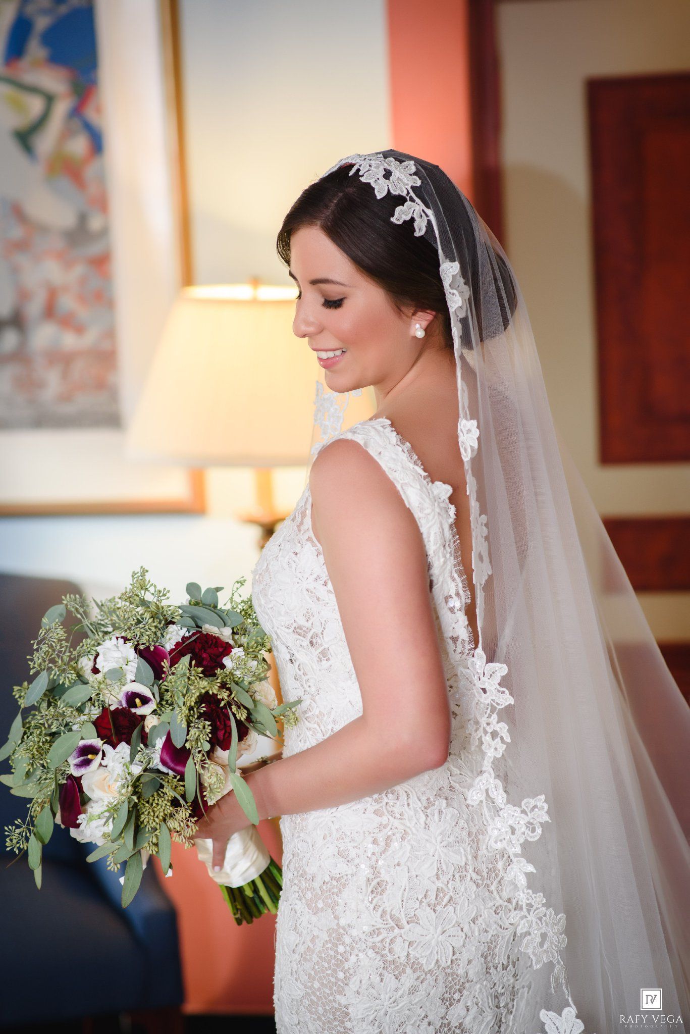 Bride in backless lace gown holding burgundy and white bouquet, wearing cathedral veil with floral headpiece.
