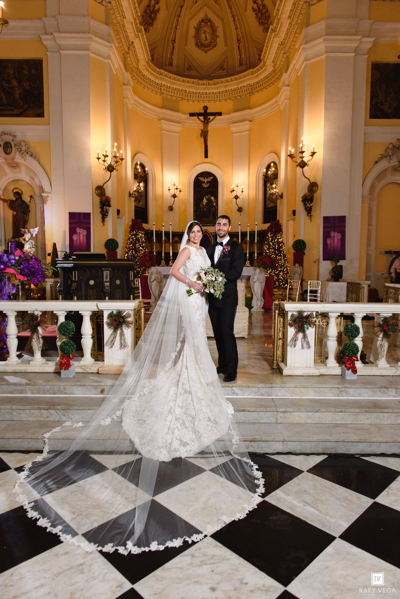 Bride and groom pose in elegant church with black and white checkered floors, ornate altar, and holiday floral decorations.