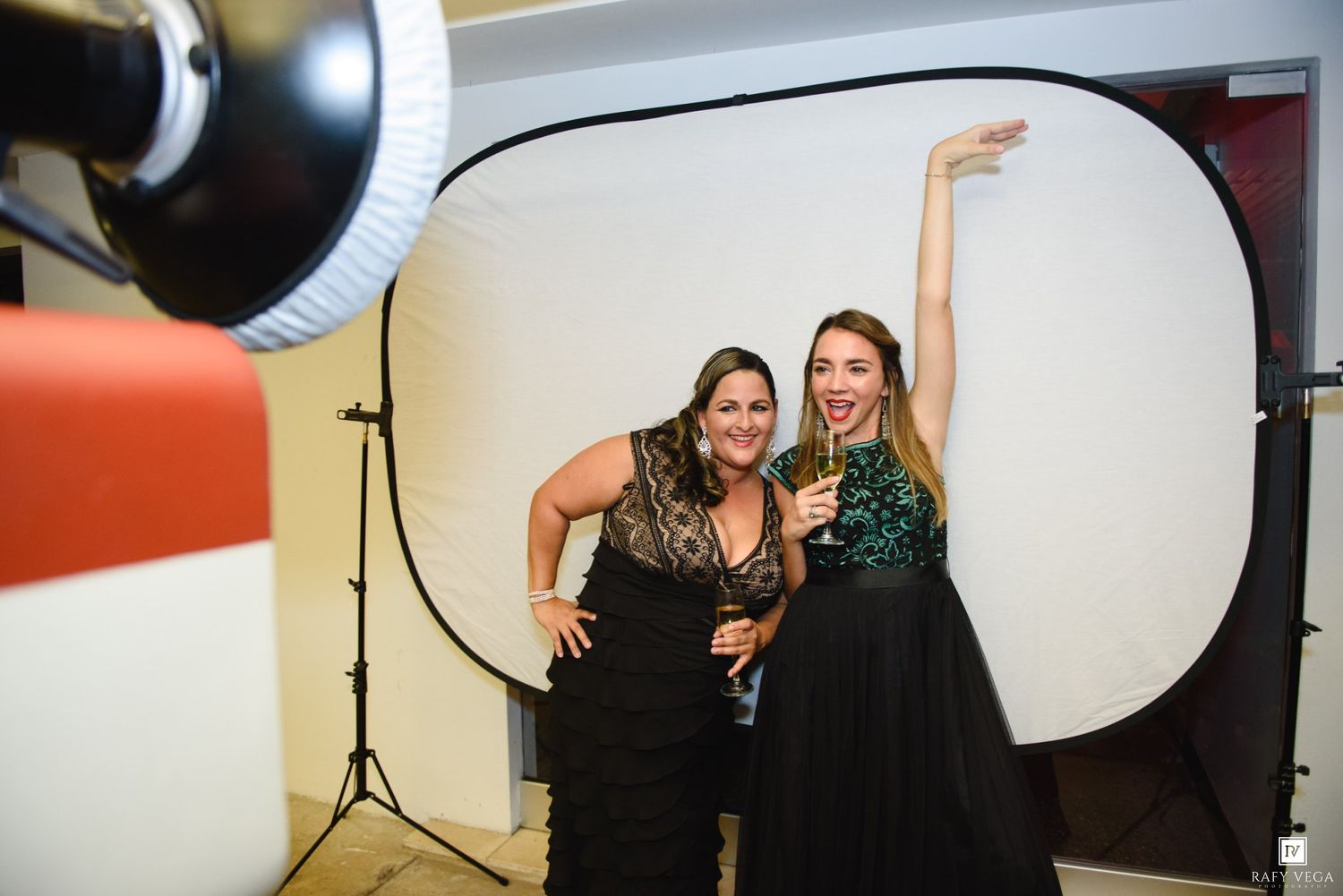 Two women in elegant gowns posing and smiling in a photo booth setup with professional lighting equipment.