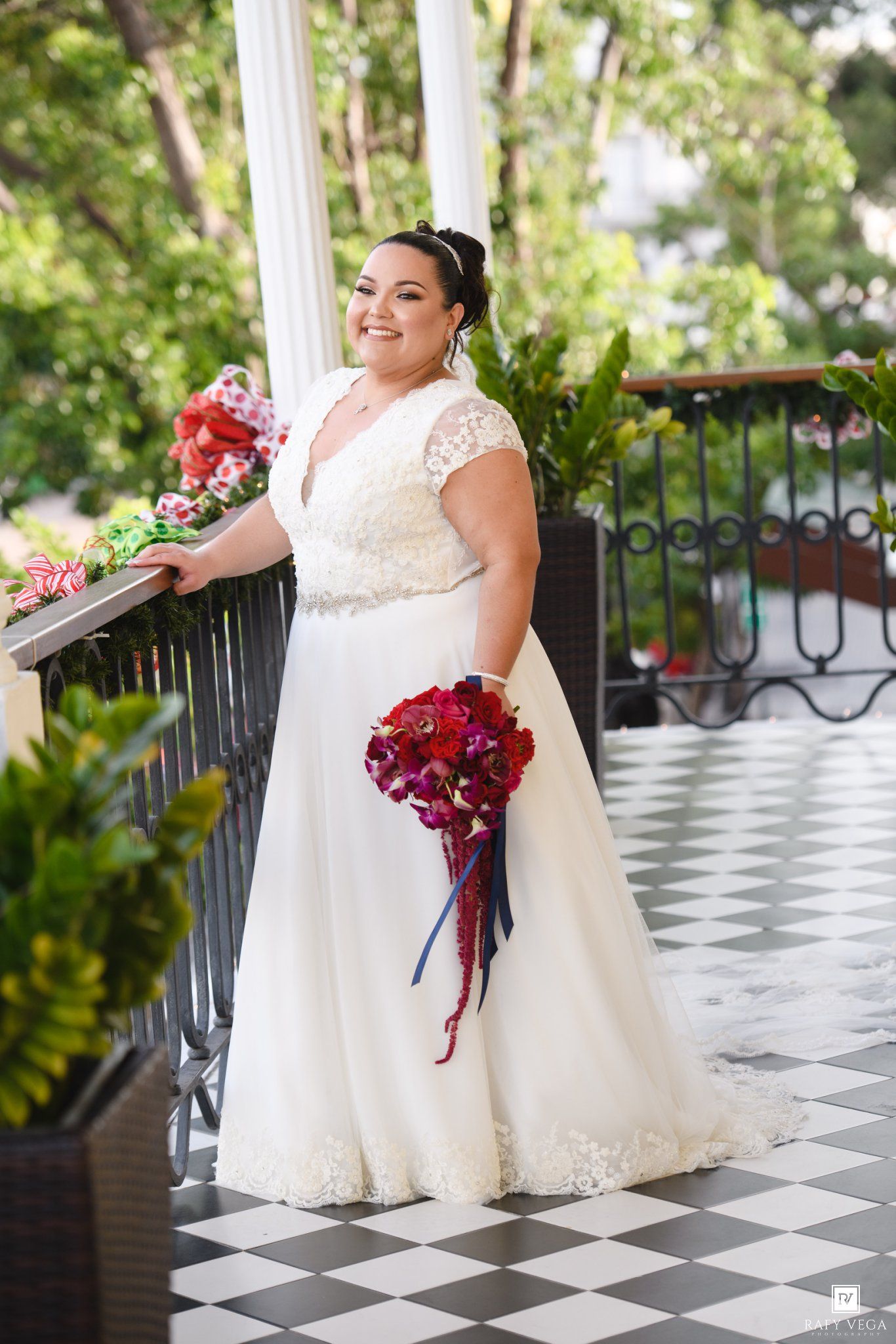 Bride in white lace V-neck wedding gown holding red bouquet on elegant balcony with black iron railing.