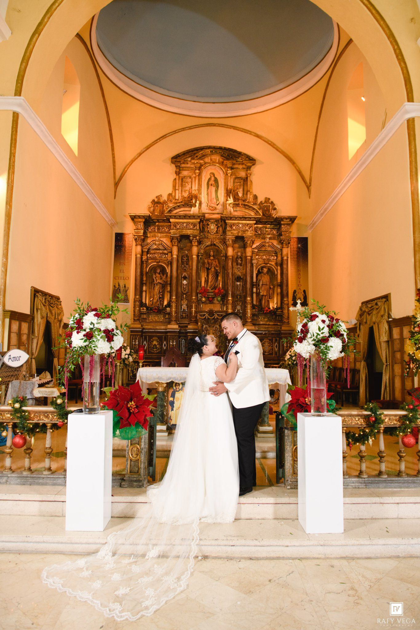 Bride and groom share a romantic moment at a church altar adorned with red floral arrangements and a golden baroque altarpiece.