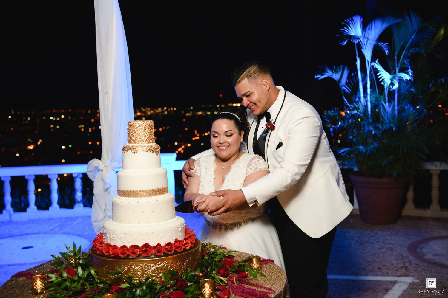 Bride and groom in white tuxedo cut tiered wedding cake on scenic night terrace with city lights.