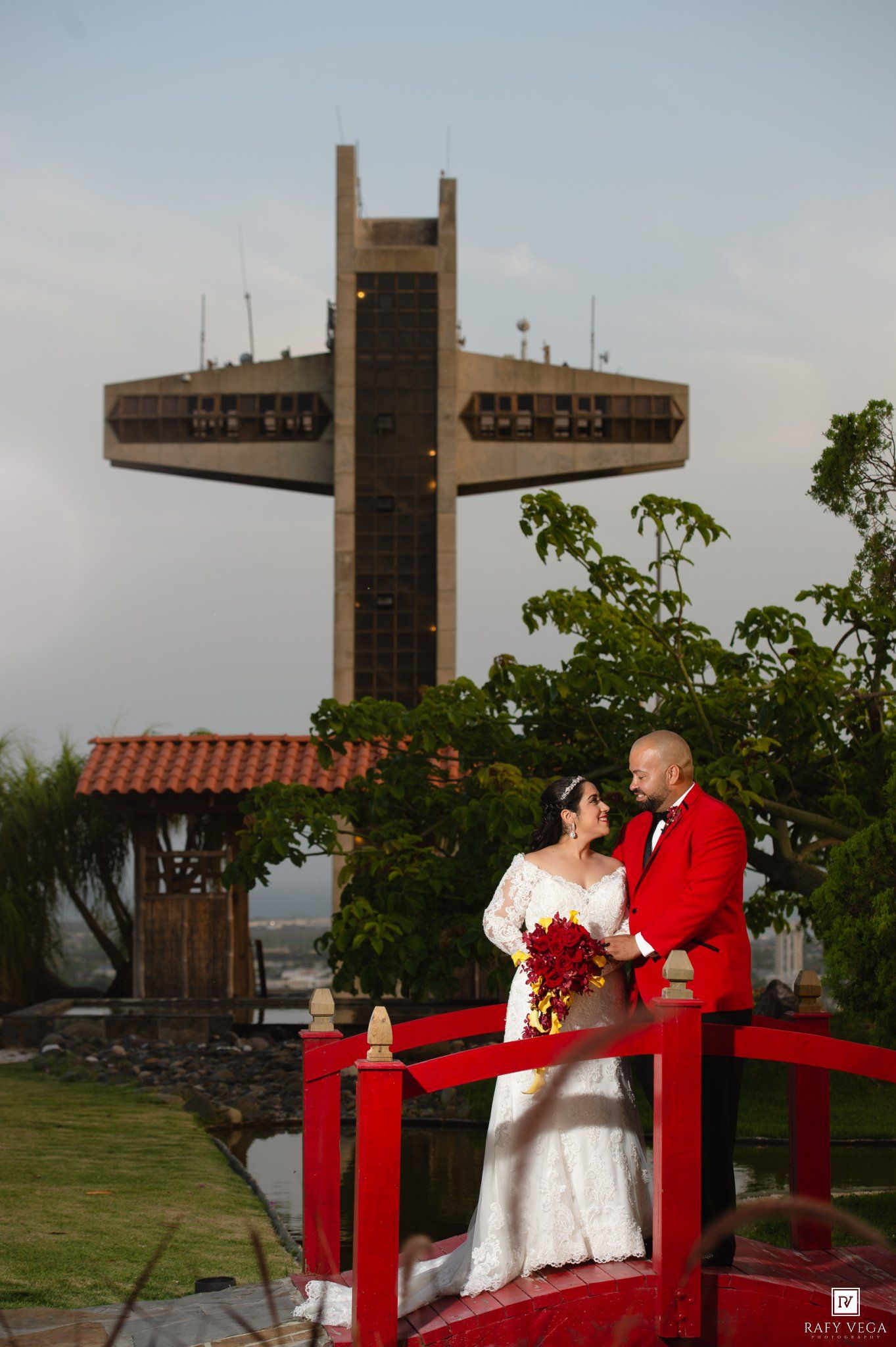 Bride and groom pose on red bridge with large cross monument behind them during outdoor wedding ceremony.