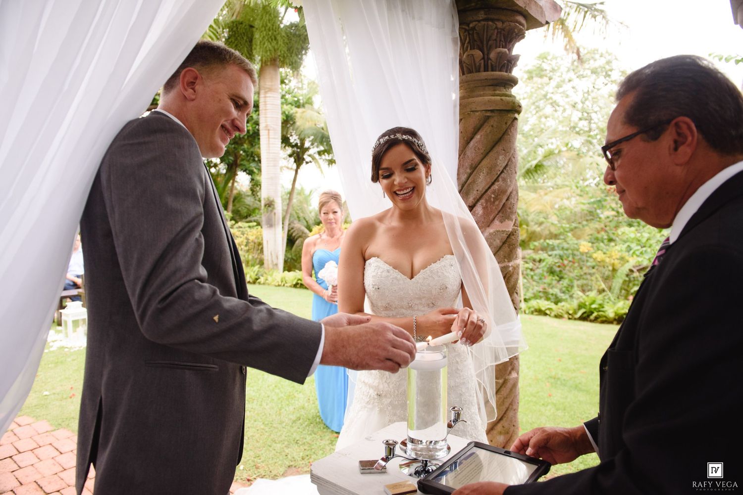 Bride and groom perform unity candle ceremony outdoors under floral arch with officiant and bridesmaid.