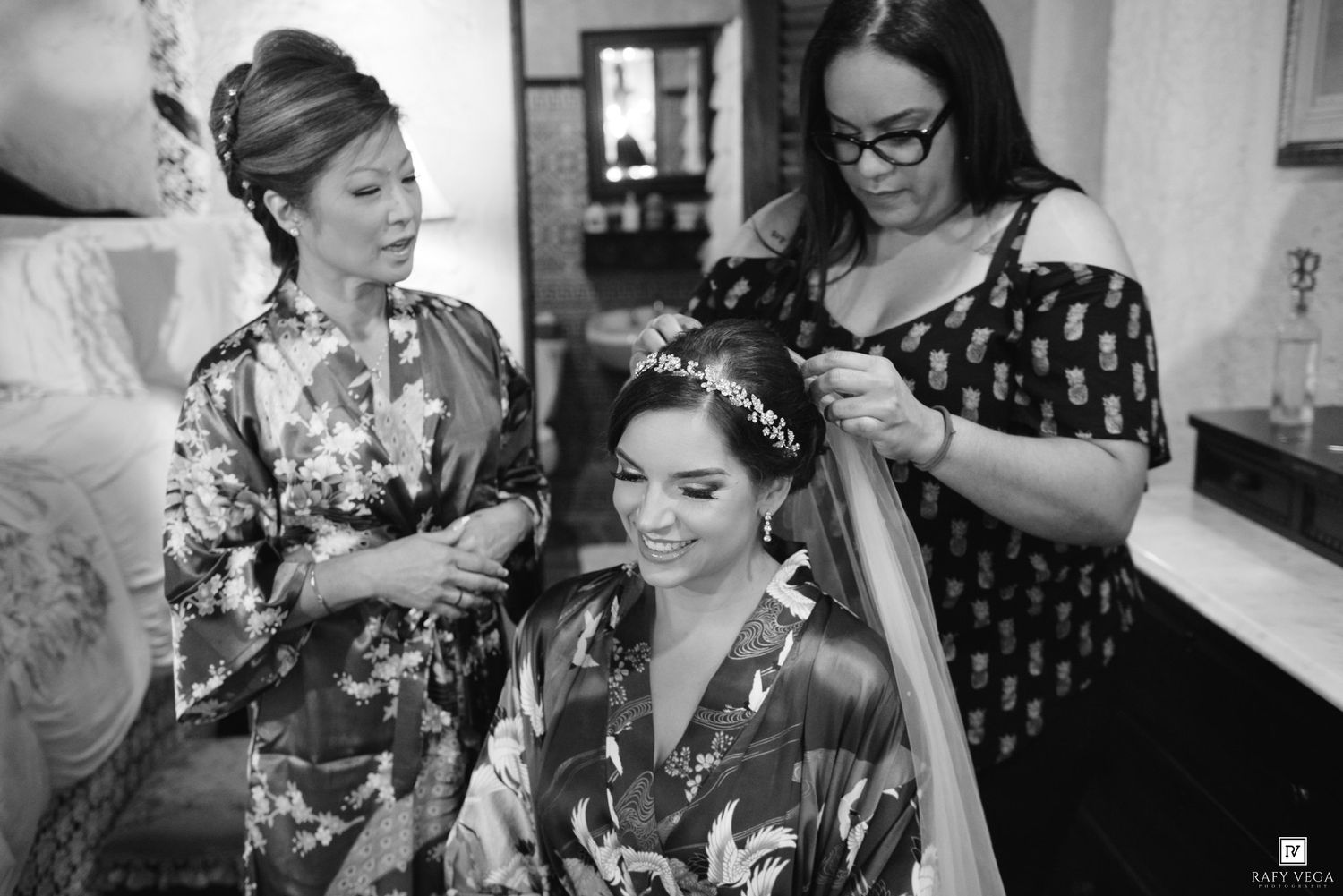 Bride smiling in floral robe as bridesmaids help style her hair and veil in a bright room.