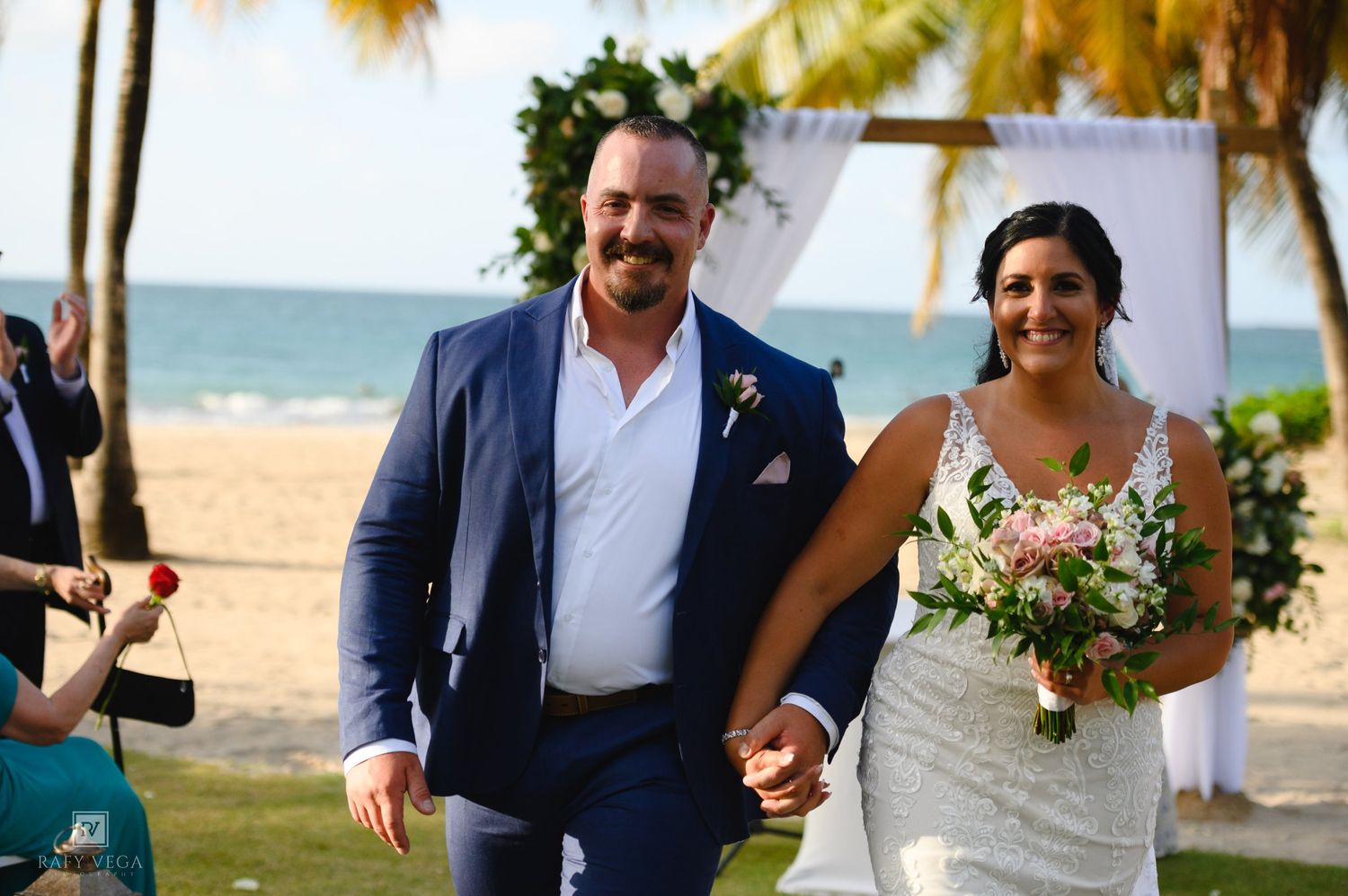 Smiling newlywed couple holding hands at a beachside wedding ceremony, groom in blue suit, bride in white gown holding bouquet.