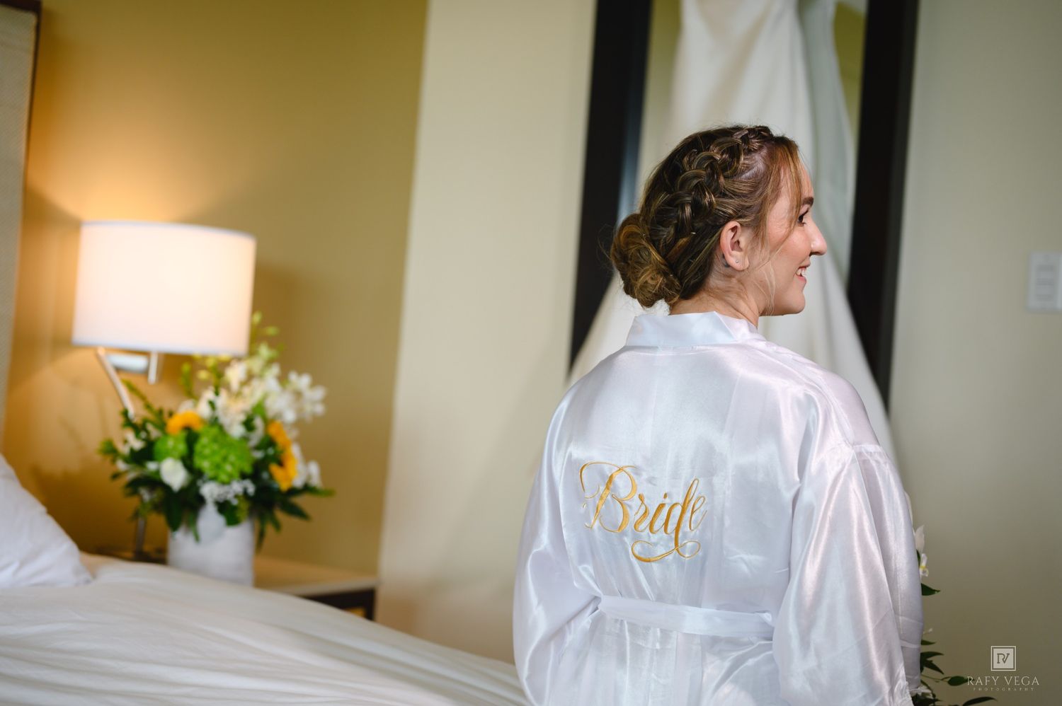 Bride wearing a white satin robe with gold embroidered script, standing in a hotel room with floral arrangements.