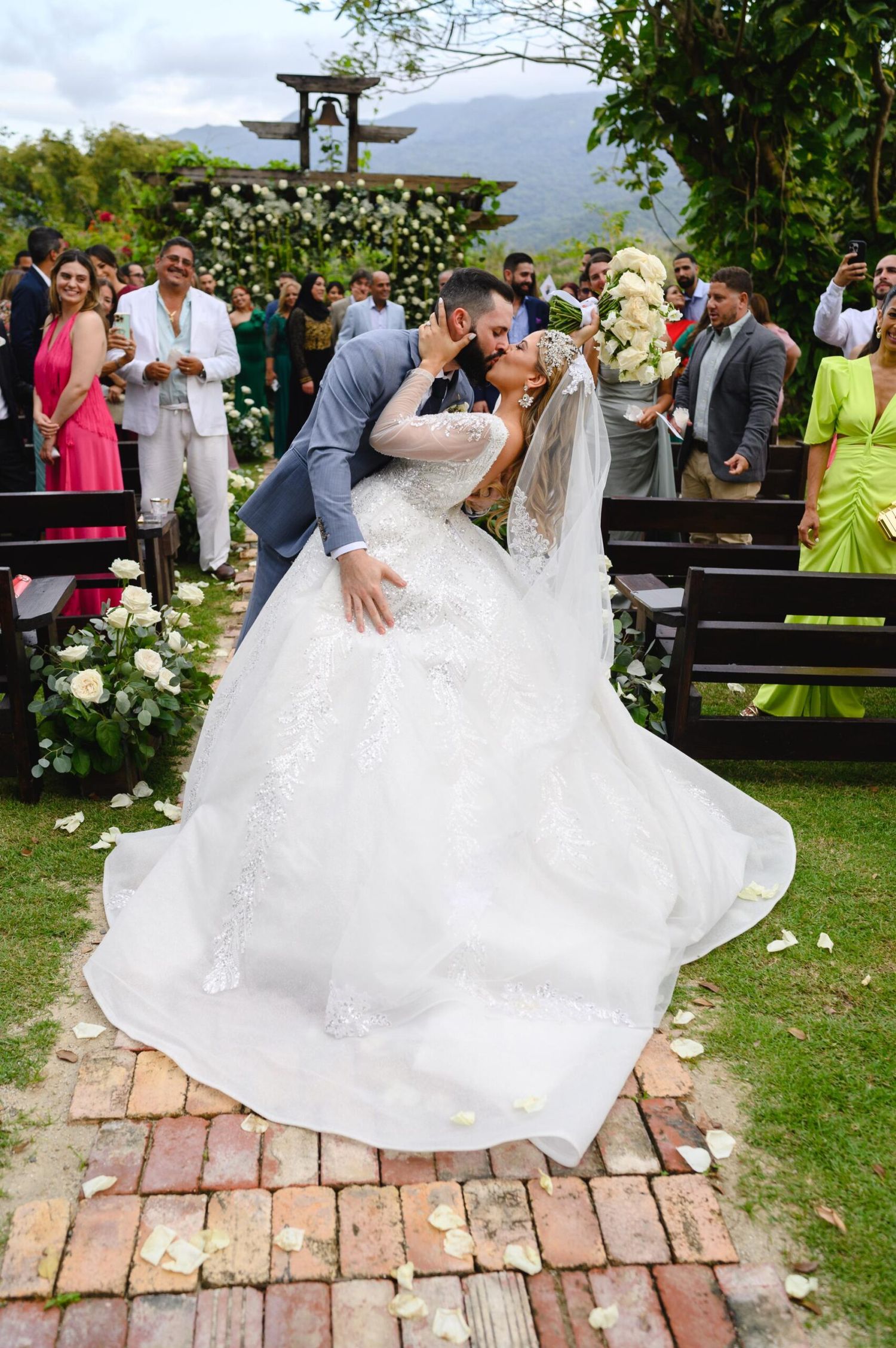 Bride and groom share a romantic kiss at outdoor wedding ceremony with guests and floral decorations.