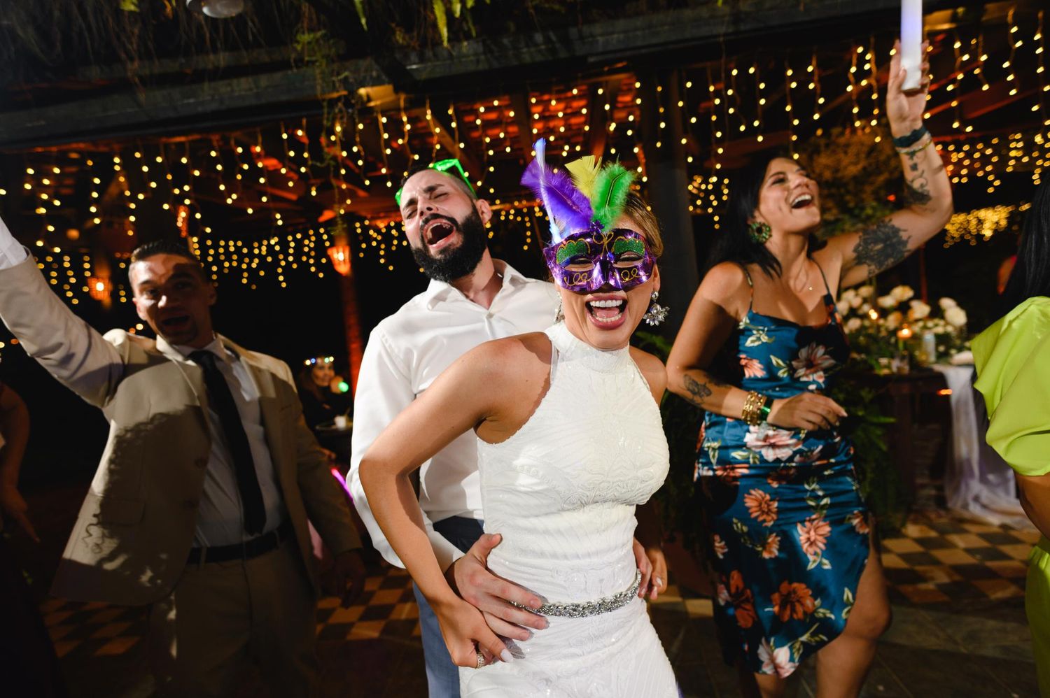 Bride and groom dancing joyfully at wedding reception, woman wearing colorful masquerade mask, festive lights in background.