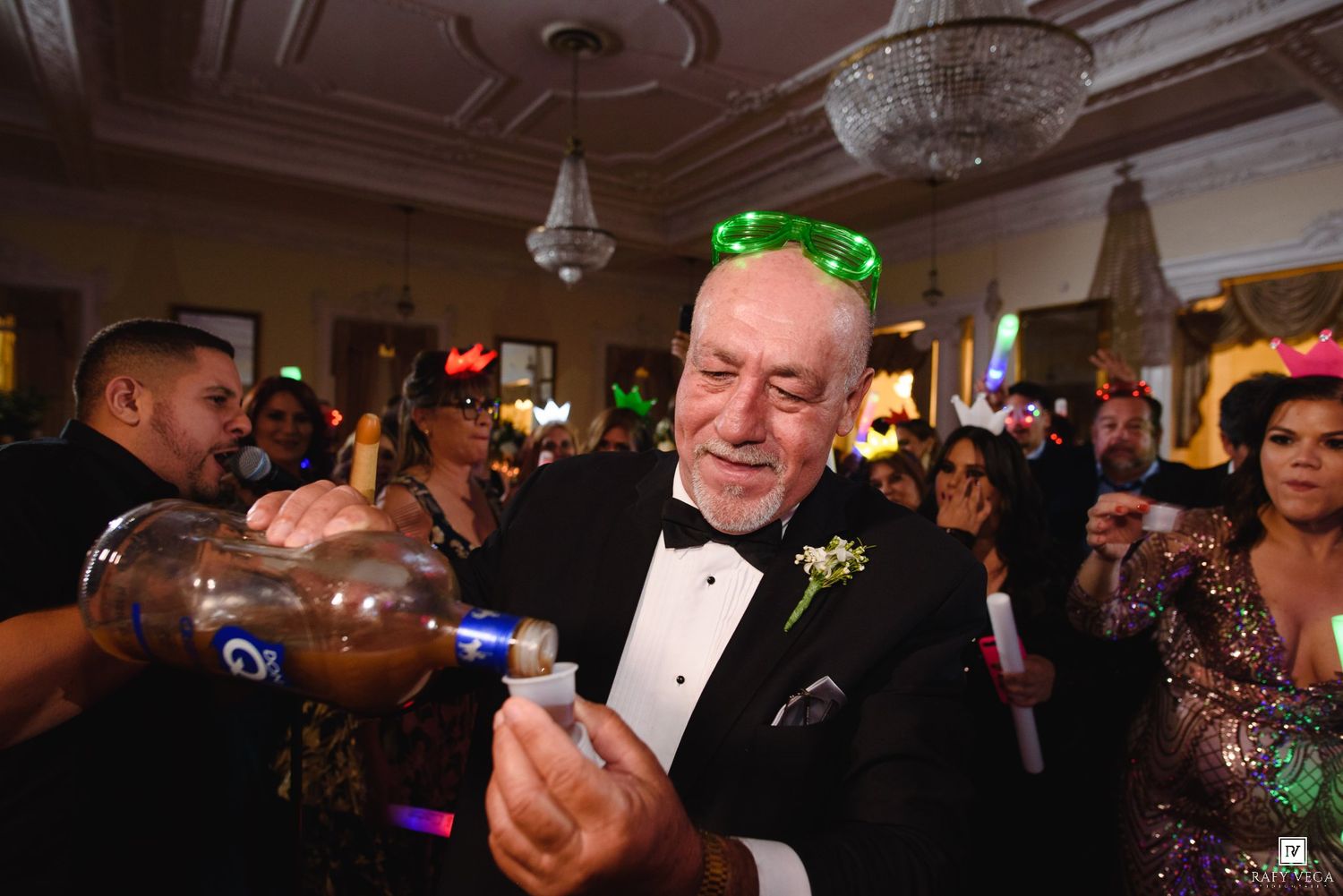 Elderly man in tuxedo pouring drinks at festive party with colorful lights and guests in formal attire.