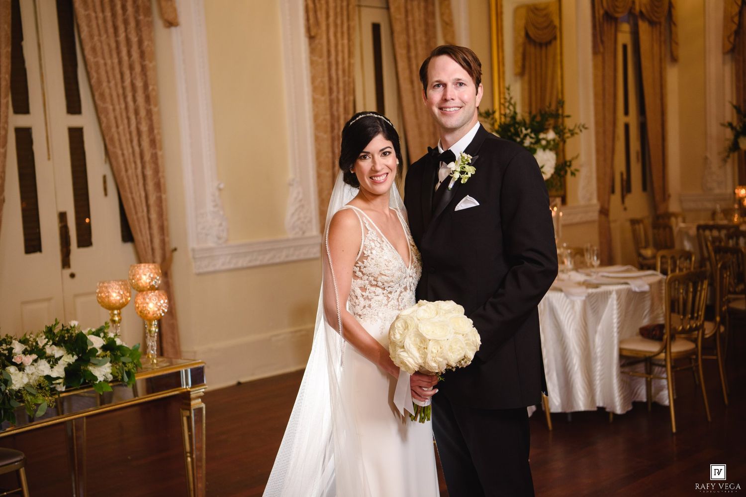 Bride and groom posing together in elegant ballroom, bride holding white bouquet in fitted gown, groom in black tuxedo.