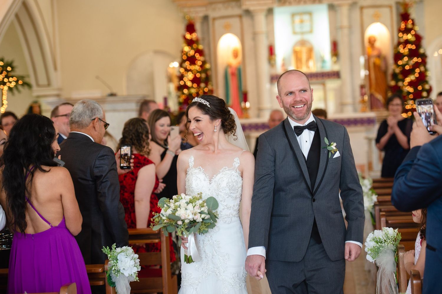 Bride and groom walk down church aisle after wedding ceremony, decorated with Christmas trees and surrounded by guests.
