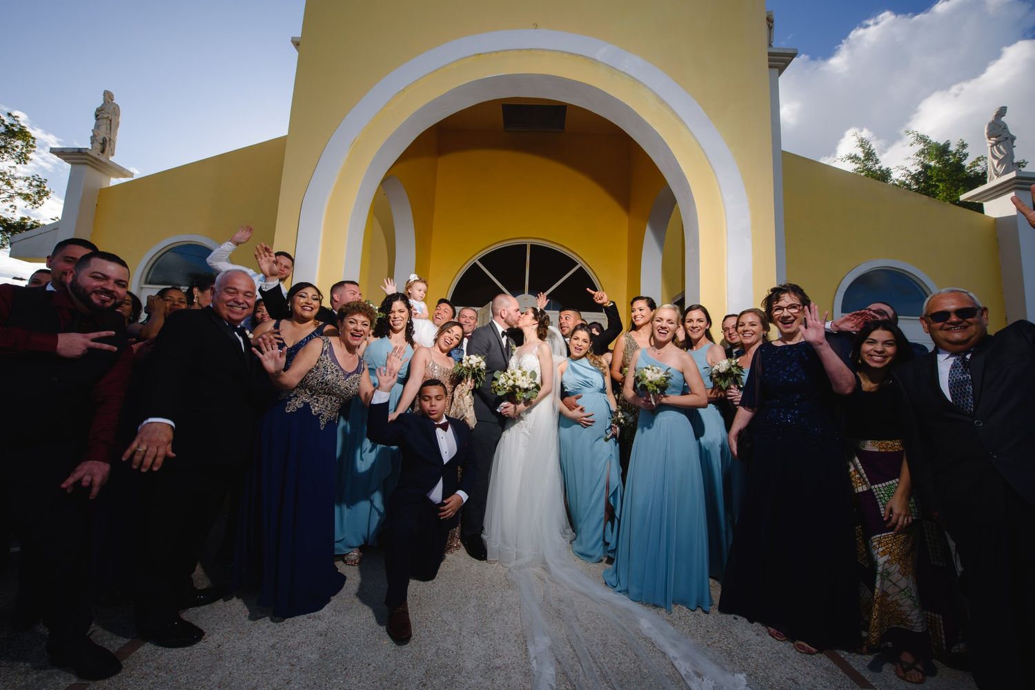 Wedding party celebrating outside a yellow chapel, bride and groom kissing surrounded by guests in blue formal attire.