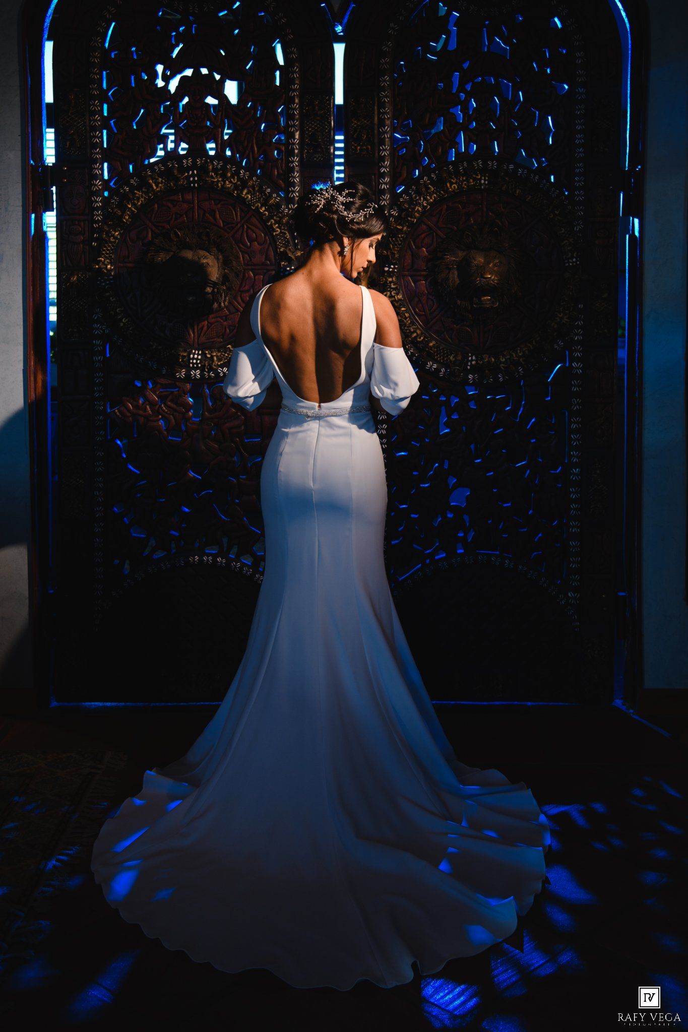 Bride in elegant backless white gown with puff sleeves stands before ornate dark doors in dramatic blue lighting.