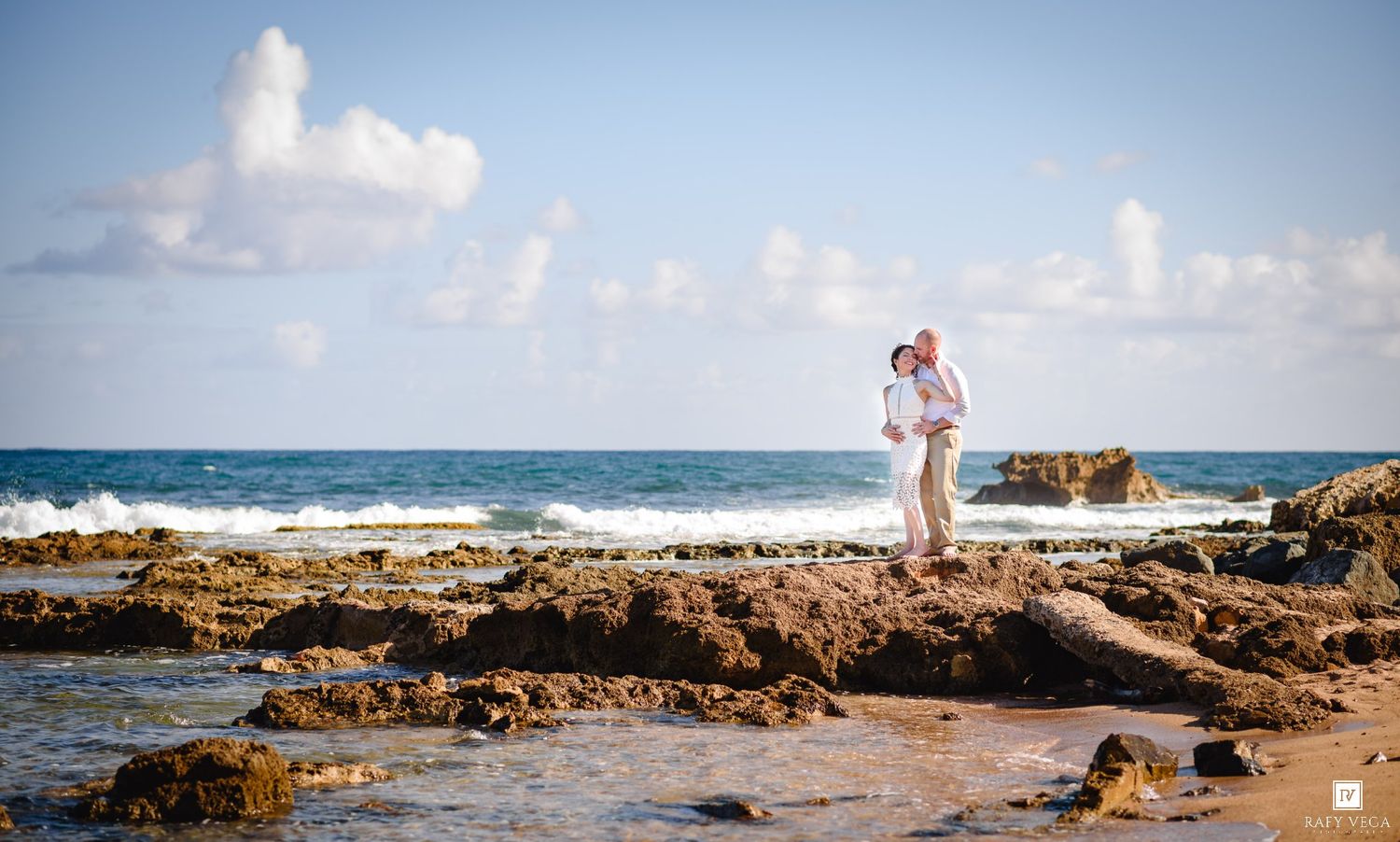Couple embracing on rocky shoreline with ocean waves and blue sky backdrop.