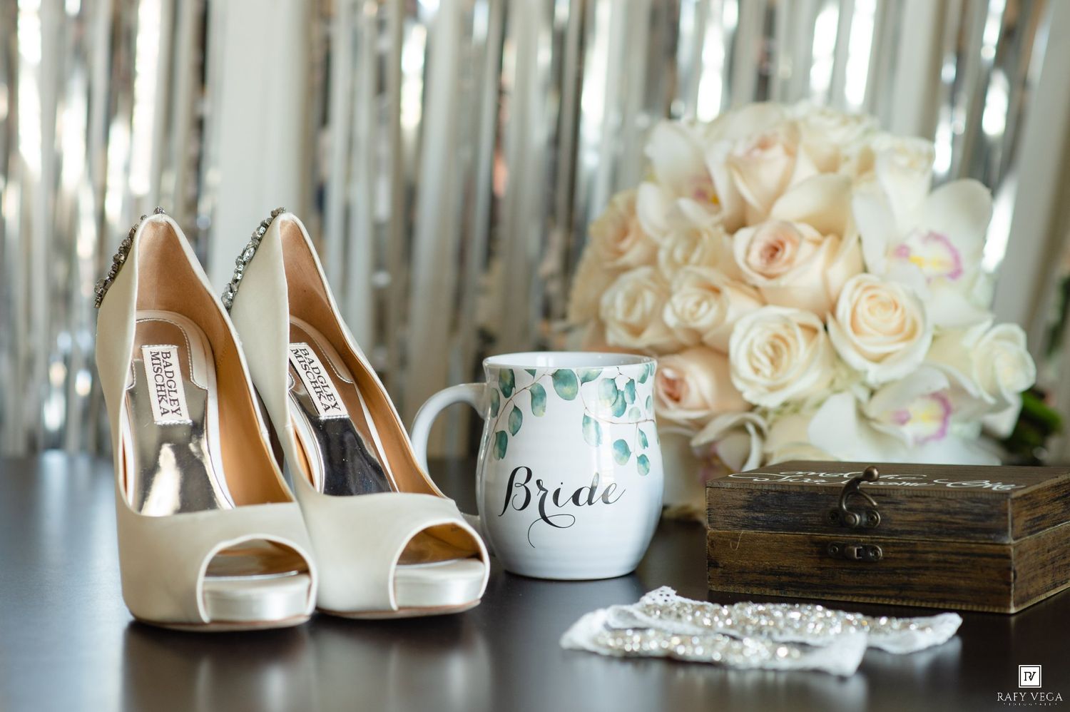 Bridal flat lay featuring Badgley Mischka heels, a Bride mug, white rose bouquet, jewelry, and a wooden box.
