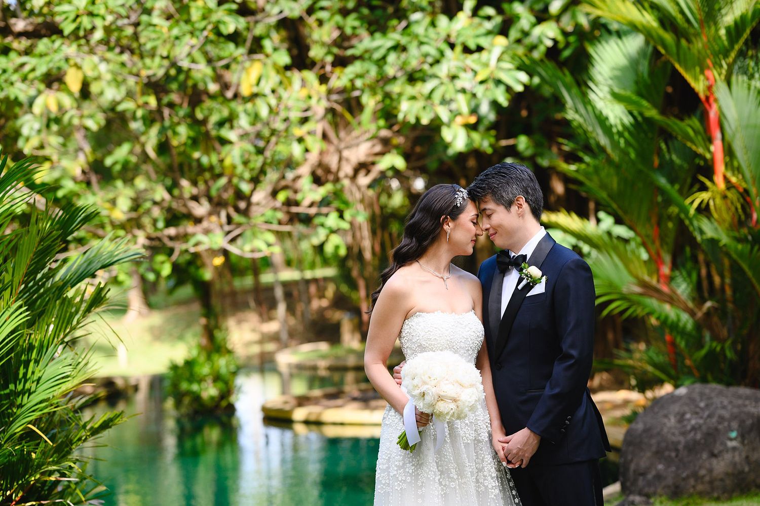 Bride and groom share a romantic kiss surrounded by lush tropical greenery and a serene blue pond on their wedding day.