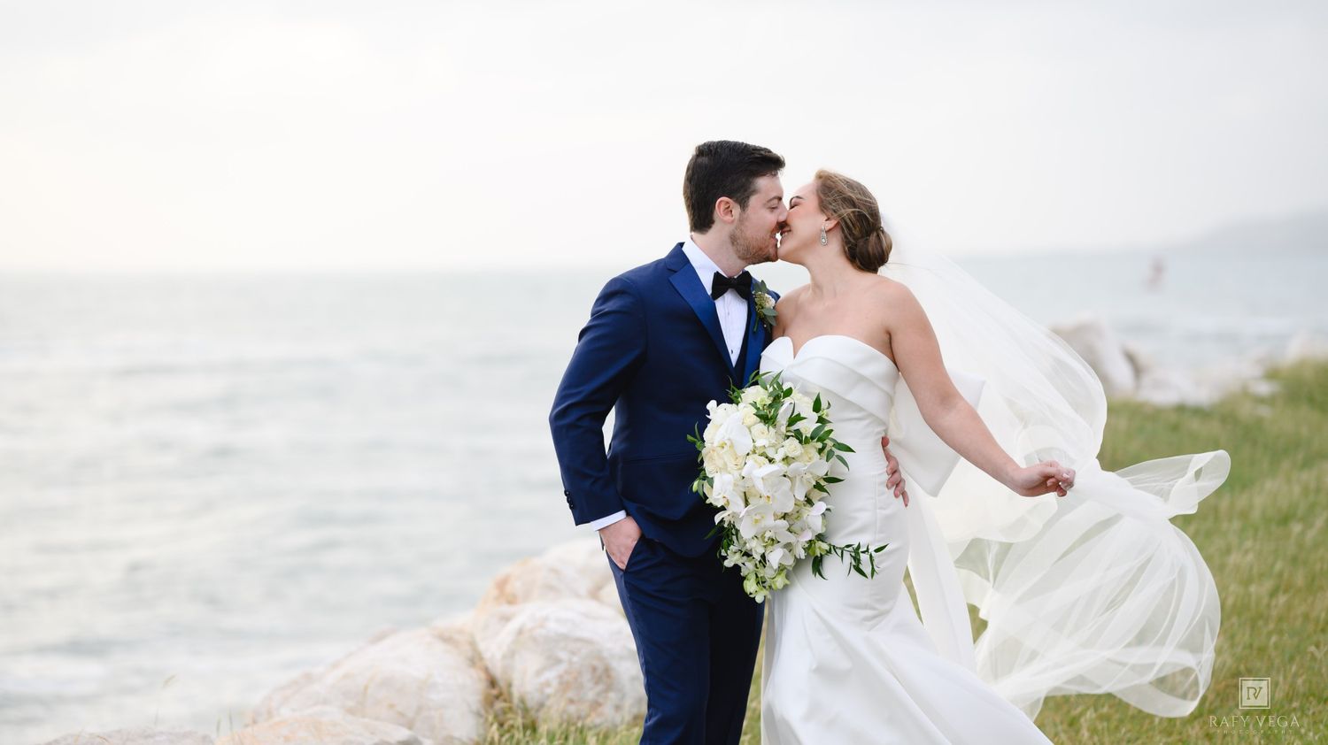 A bride and groom share a romantic kiss by the ocean, her veil flowing in the wind, as she holds a white bouquet.