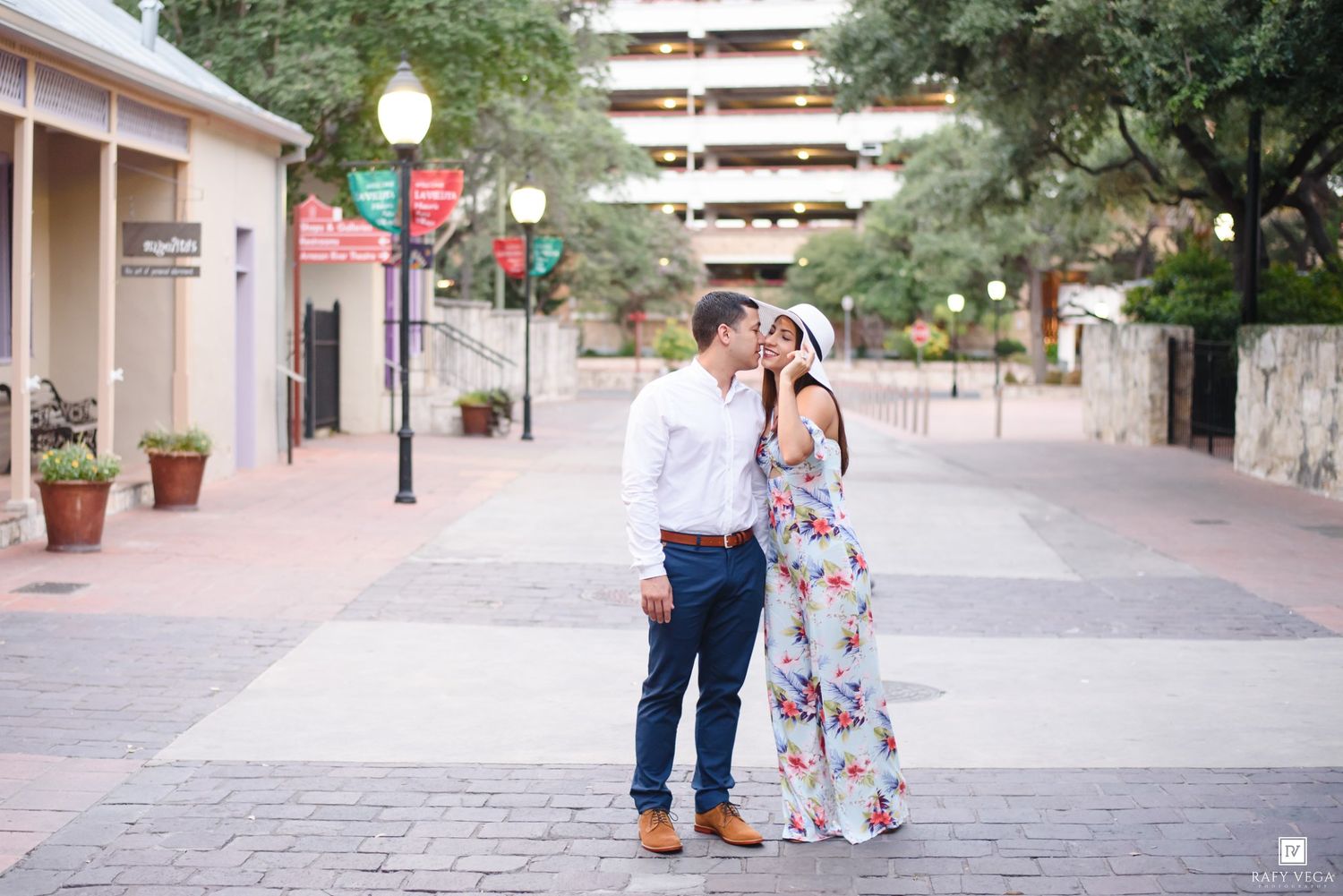 Couple sharing a romantic kiss on a charming outdoor plaza with string lights and lush greenery in the background.