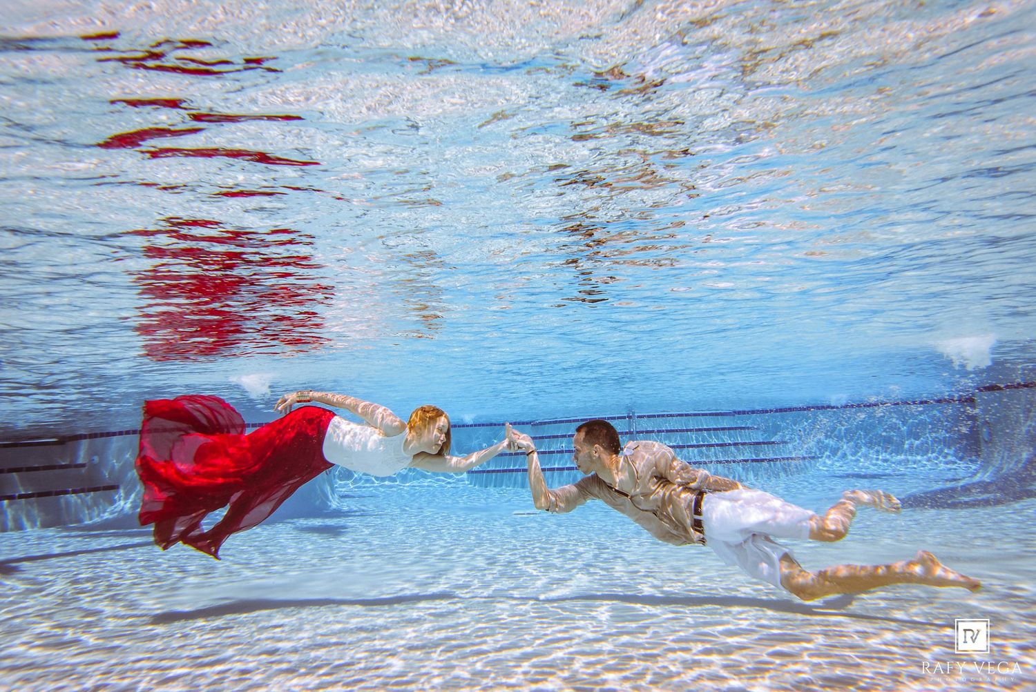 Underwater wedding photo of bride in red gown and groom in white suit swimming gracefully in a pool.
