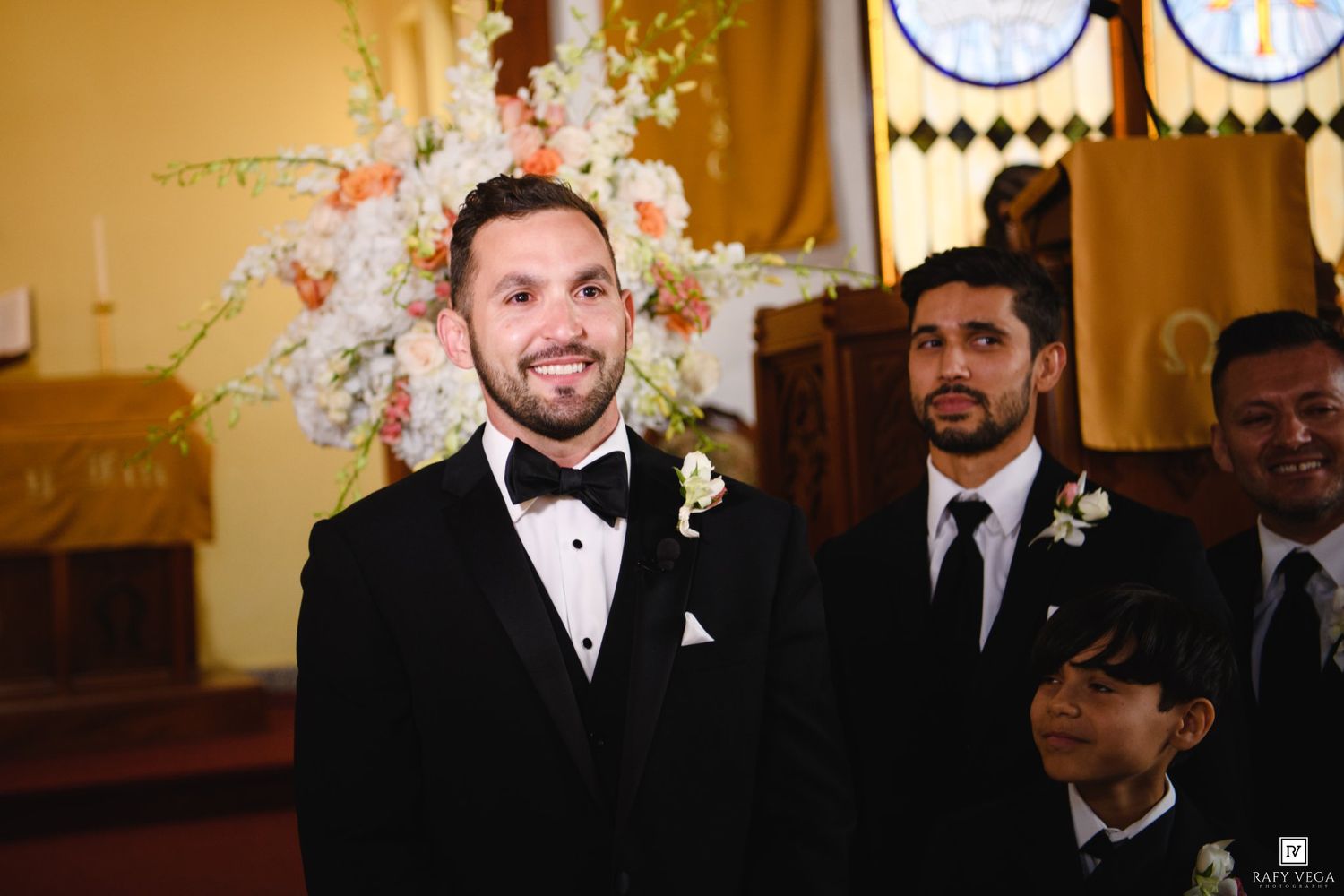 Groom smiling at altar in black tuxedo with boutonniere, groomsmen standing behind in church wedding ceremony.