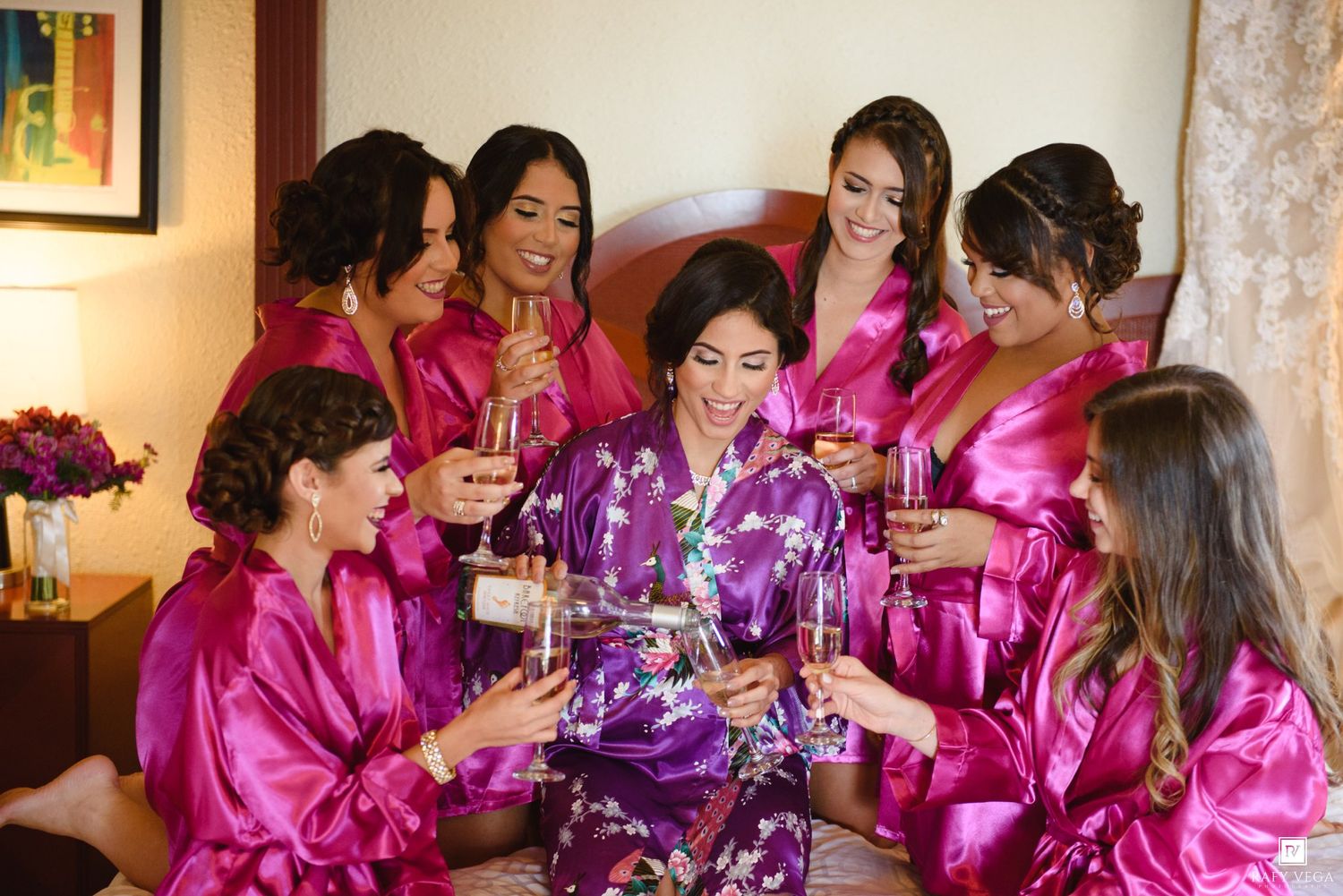 Bride and bridesmaids in pink satin robes toasting with champagne glasses during wedding morning preparations.