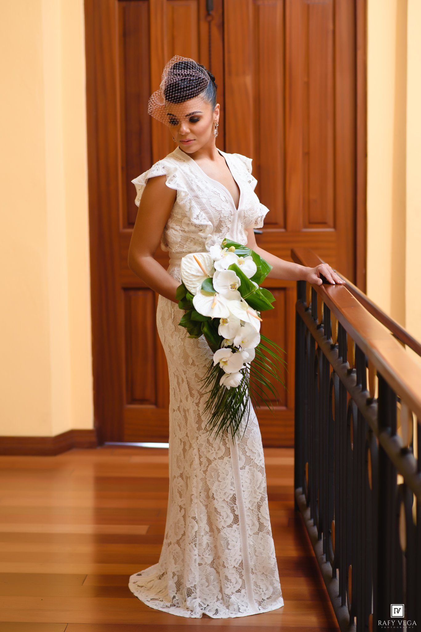 Elegant bride in white lace gown holding cascading calla lily bouquet, standing by railing in grand hall.