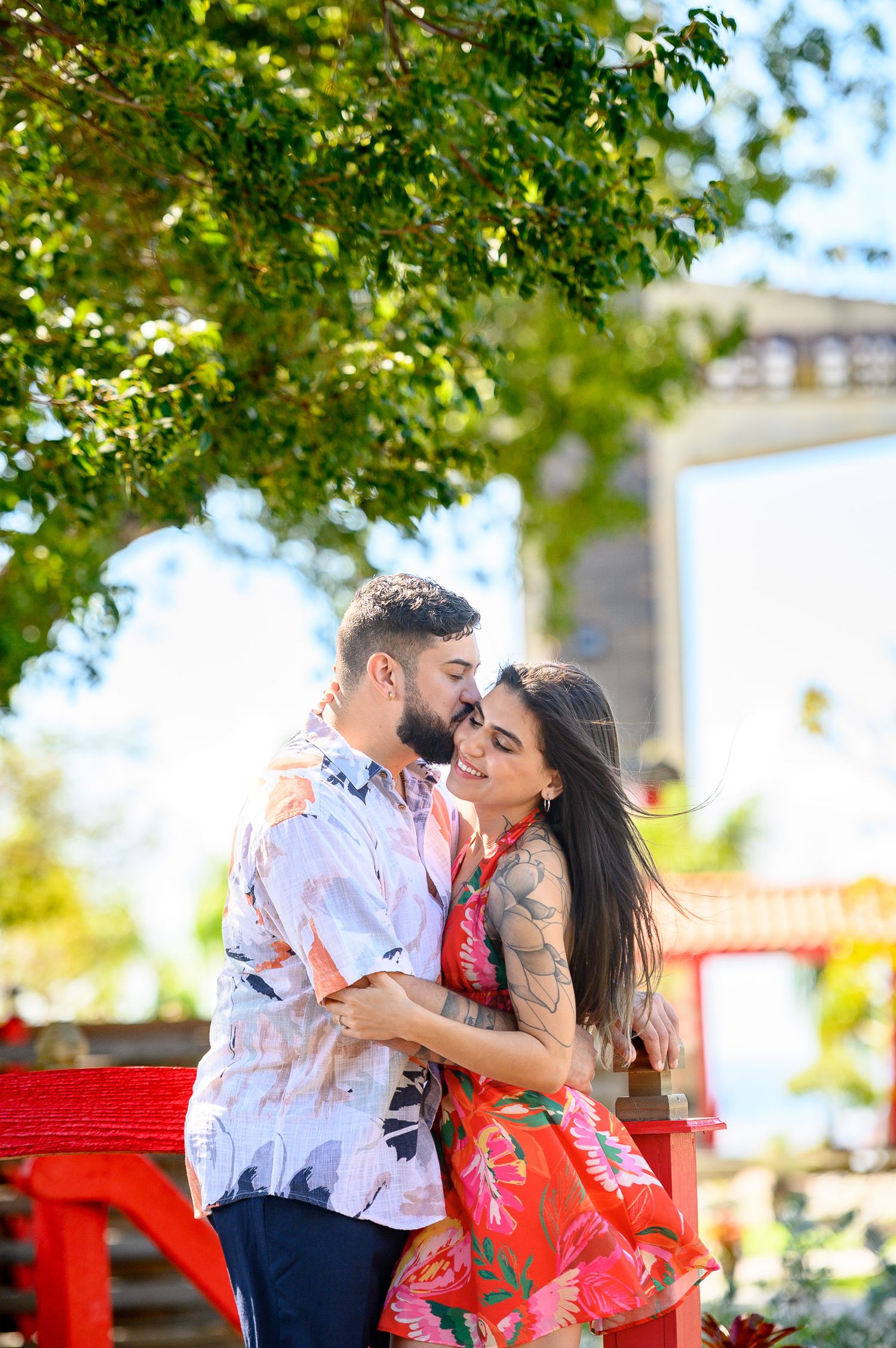 A couple in floral outfits share a romantic moment outdoors, surrounded by lush green trees and vibrant red decor.