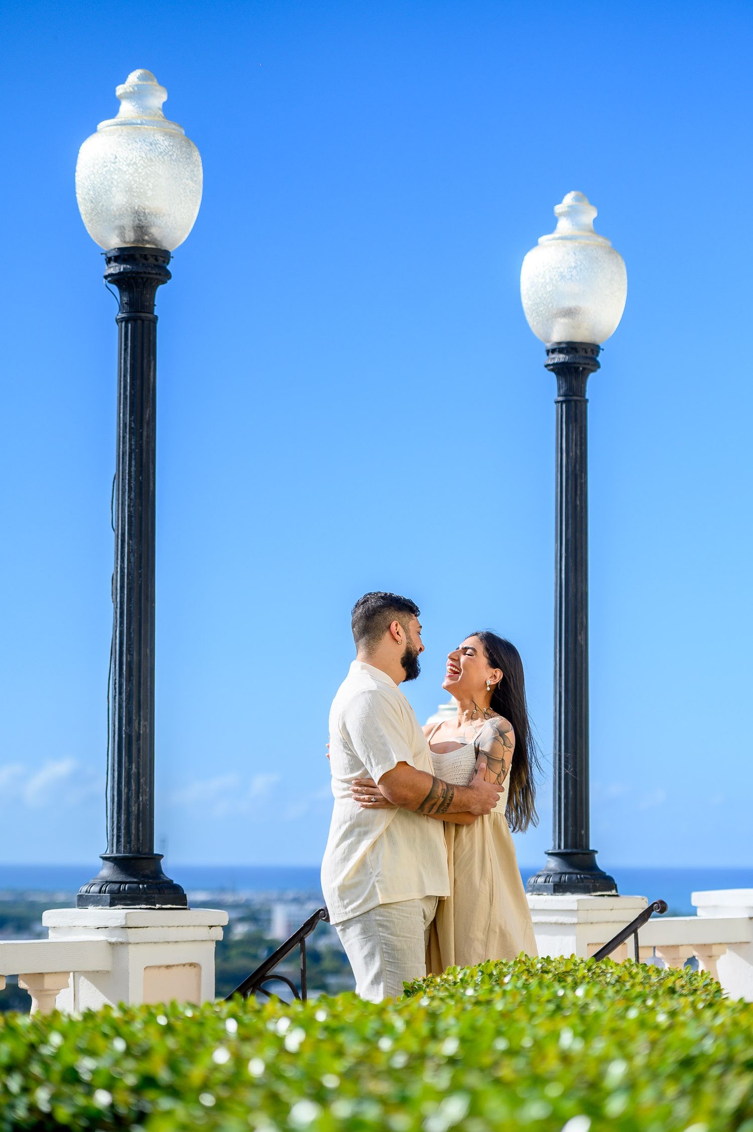 Couple embracing between ornate lamp posts under a clear blue sky, wearing matching cream outfits in an outdoor setting.