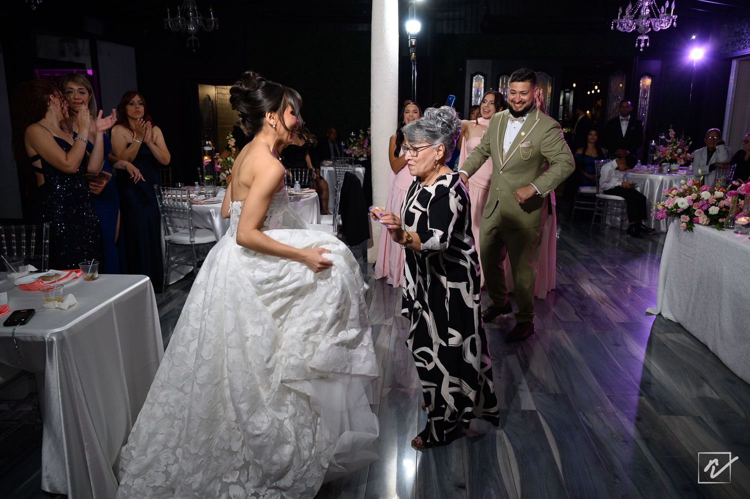 Bride in a white ball gown dances with an elderly woman in a floral dress at a wedding reception while guests watch nearby.