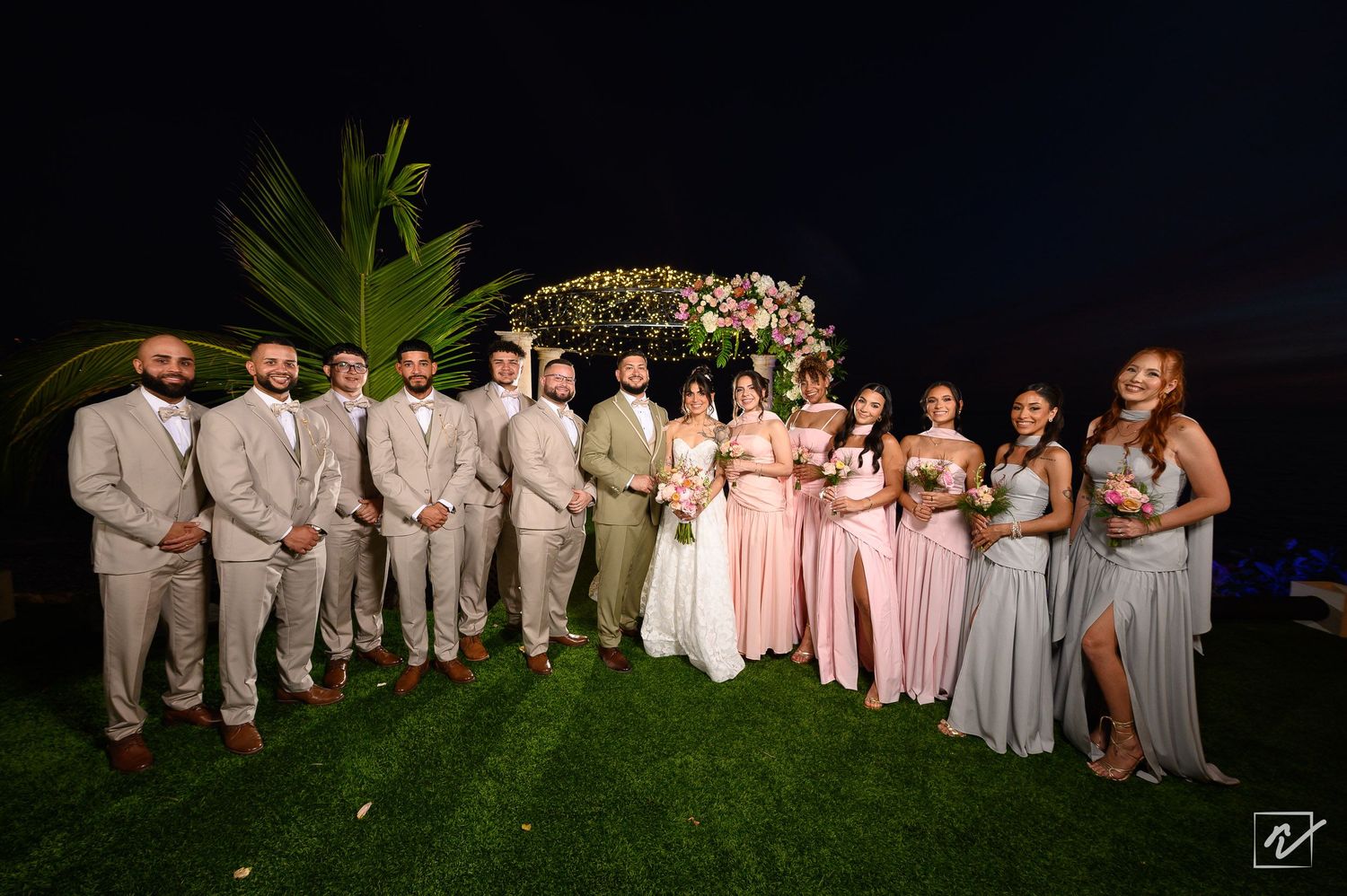 Bride and groom pose with groomsmen in tan suits and bridesmaids in pink and gray dresses on a lawn at a night outdoor wedding.