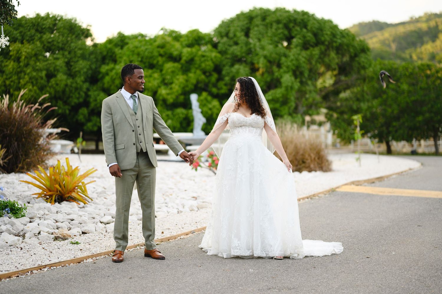 Interracial couple in wedding attire holding hands outdoors, bride in white gown, groom in gray suit, green hills behind.
