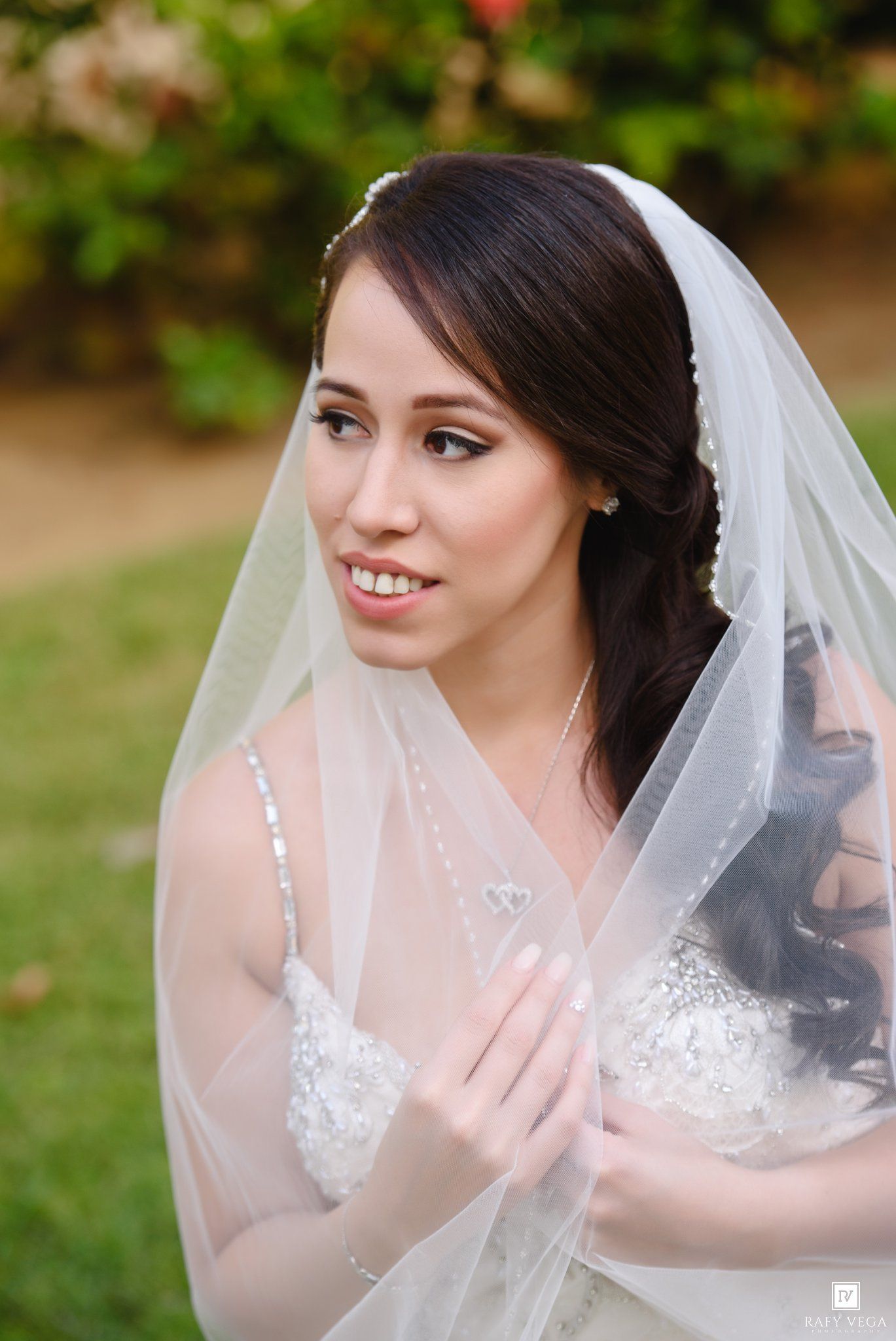 A beautiful bride in a white lace gown and veil smiles softly outdoors, with greenery in the background.