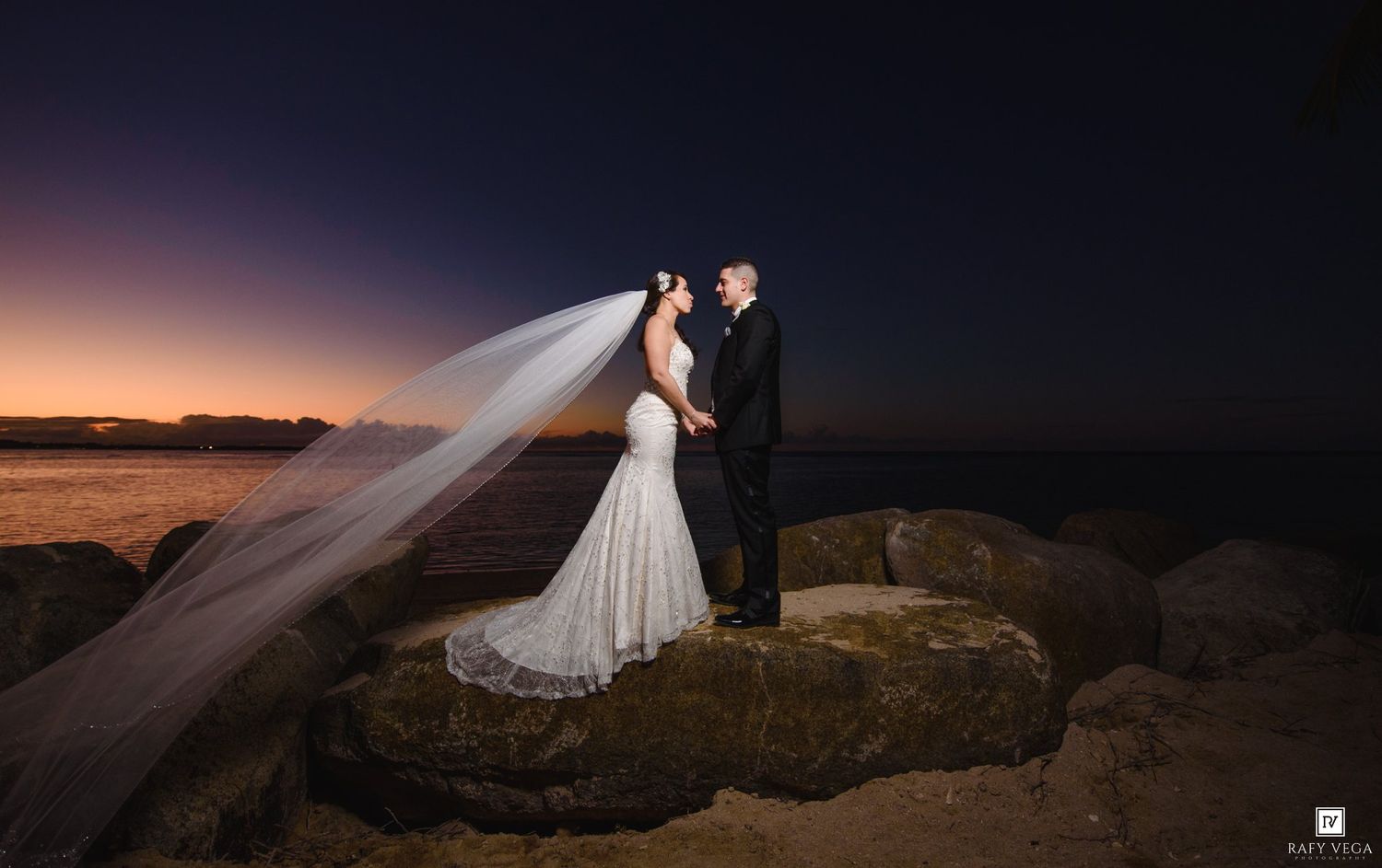 Bride and groom share romantic kiss on rocky shore at sunset, wedding veil flowing in the breeze.