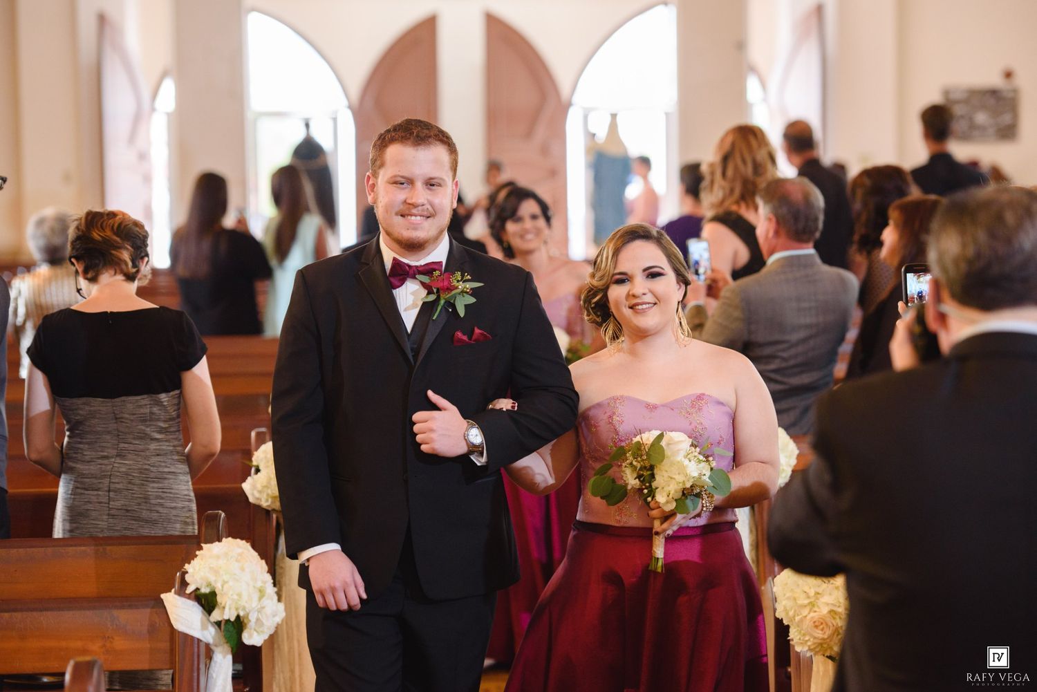 Groomsman and bridesmaid in burgundy dress walk down church aisle together during wedding ceremony.
