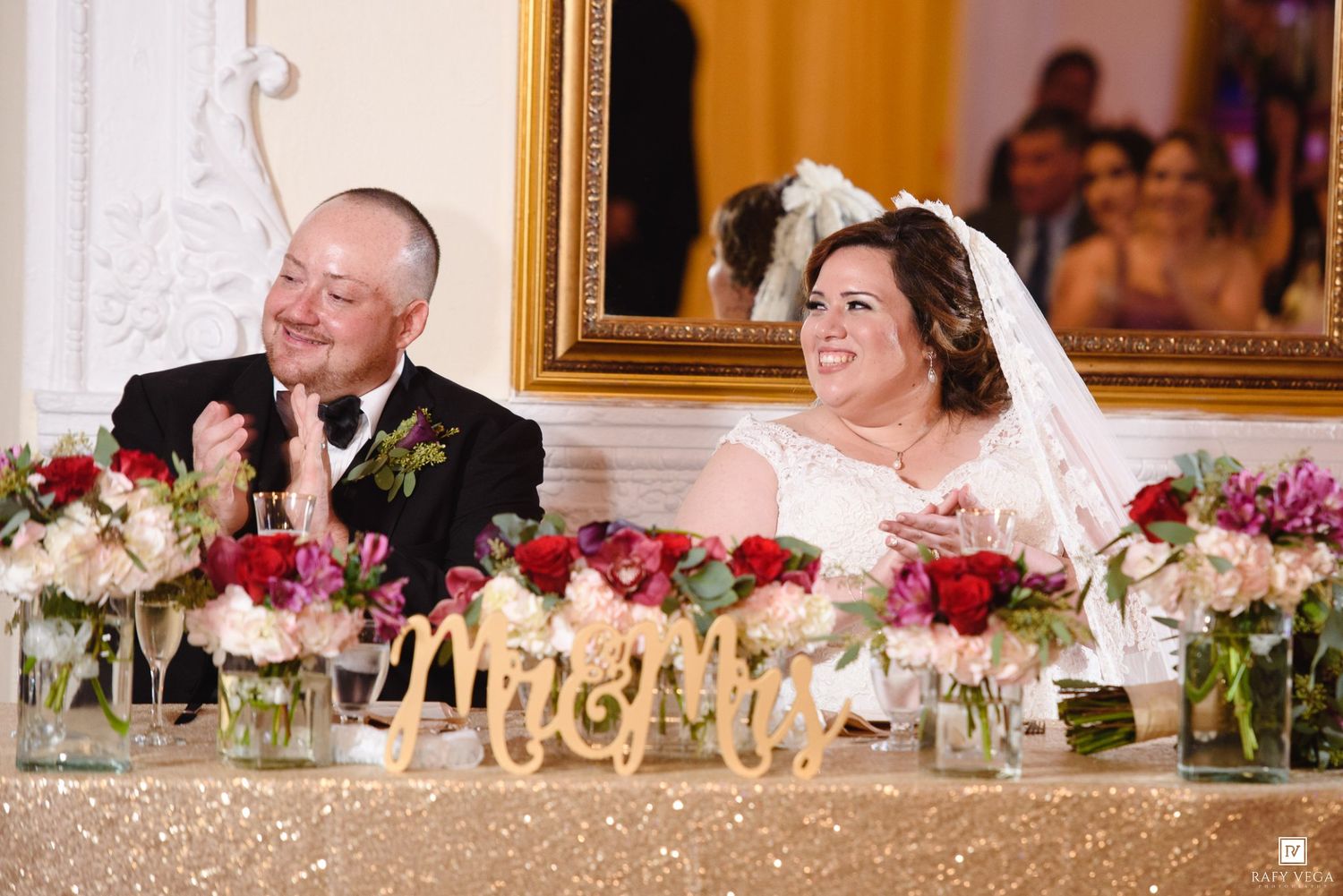 Bride and groom laughing at wedding reception head table with Mr & Mrs sign and floral arrangements.