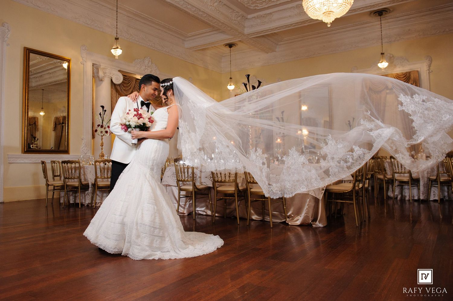 Bride and groom share romantic kiss in elegant ballroom, her veil dramatically flowing in the air during wedding reception.