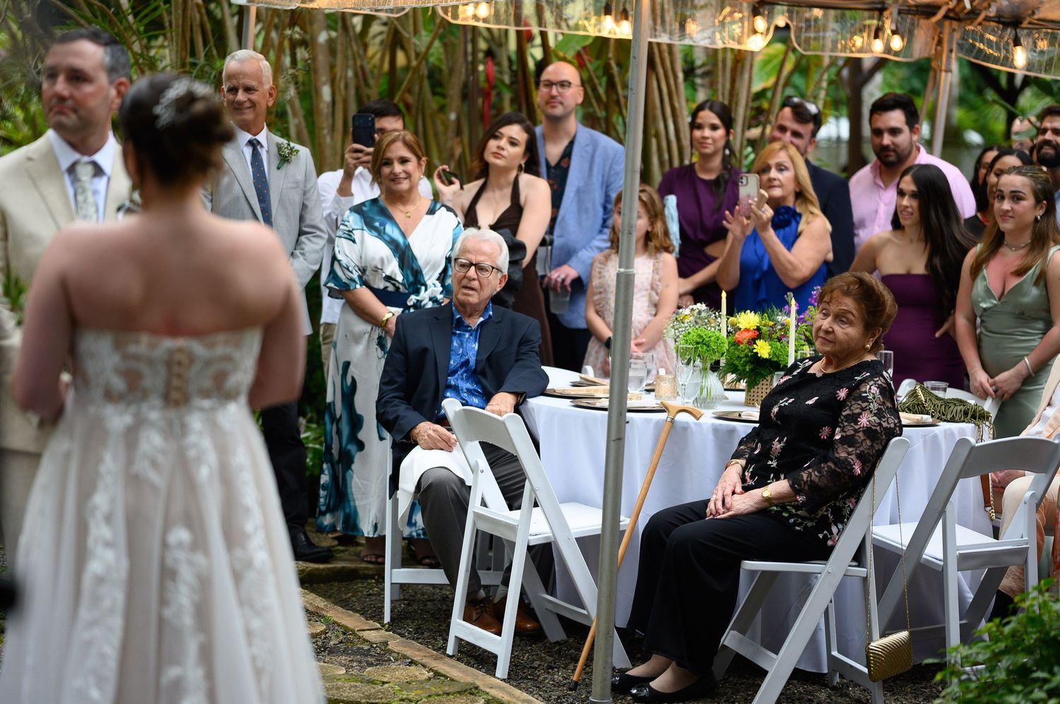 Guests watch a bride in a white lace gown at an outdoor wedding ceremony surrounded by lush greenery.