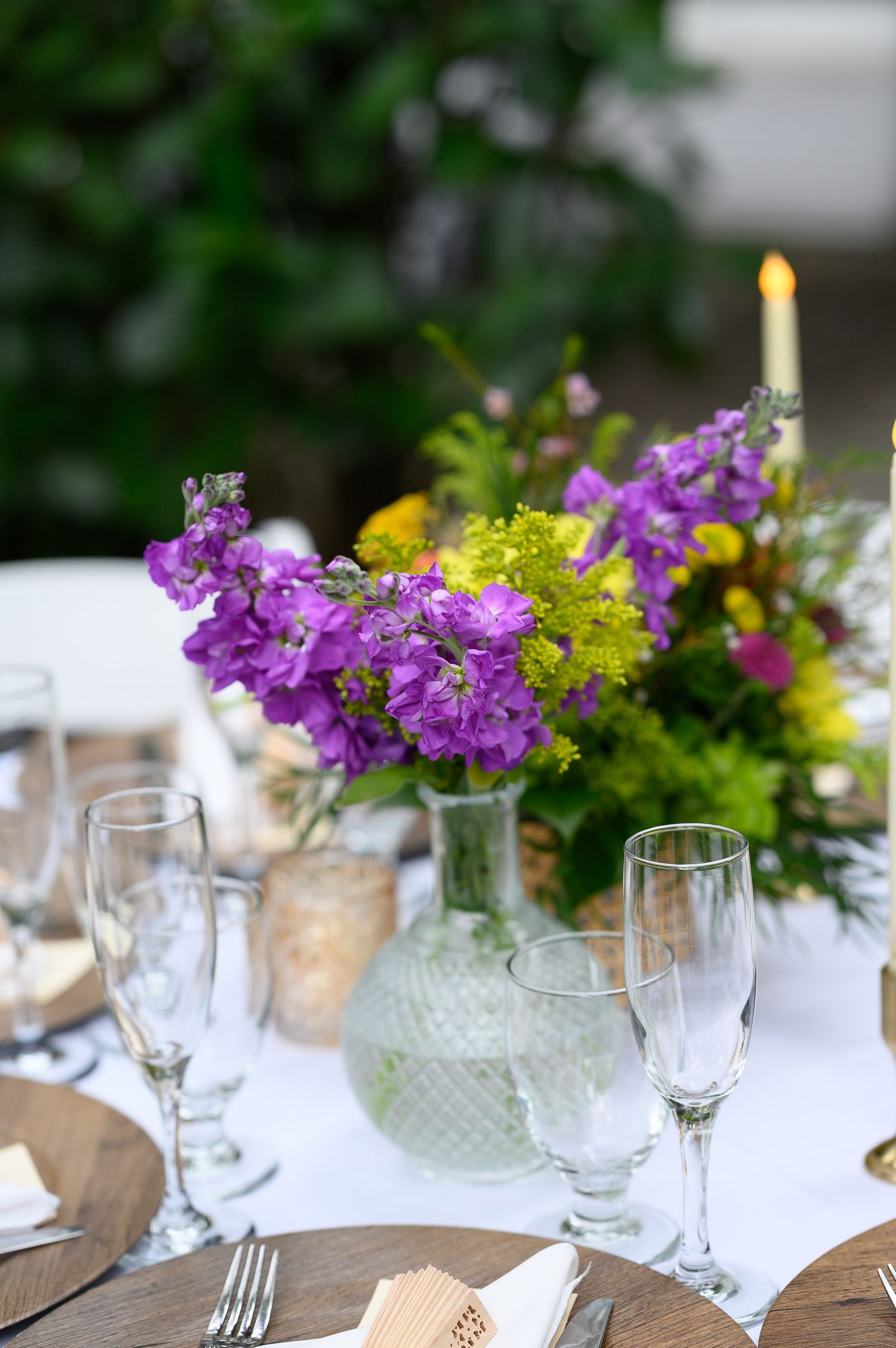 Elegant outdoor table setting with purple flowers, crystal glasses, and a candle centerpiece on a white tablecloth.