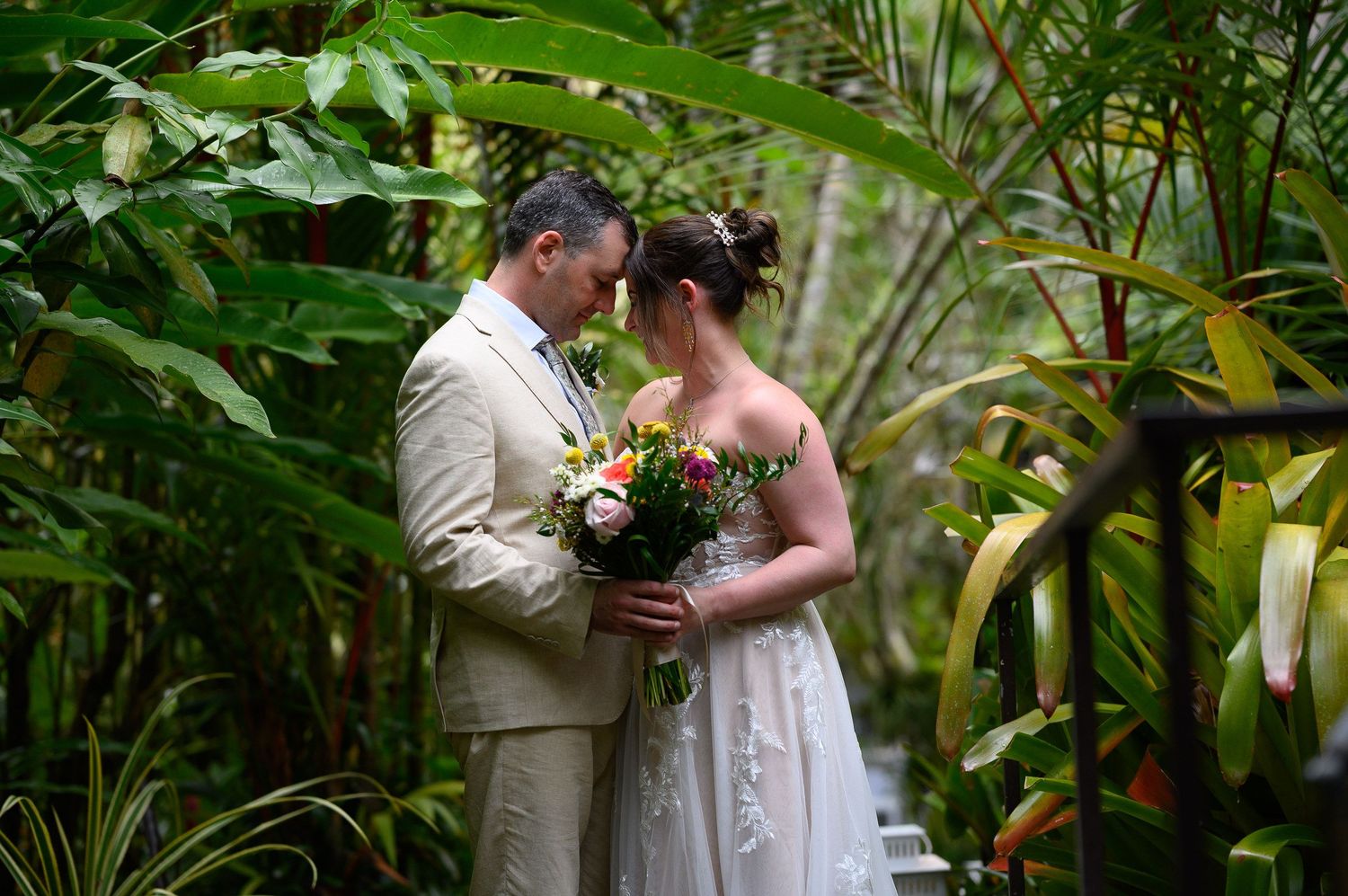 Bride and groom share an intimate moment surrounded by lush tropical greenery, bride holding a colorful bouquet.