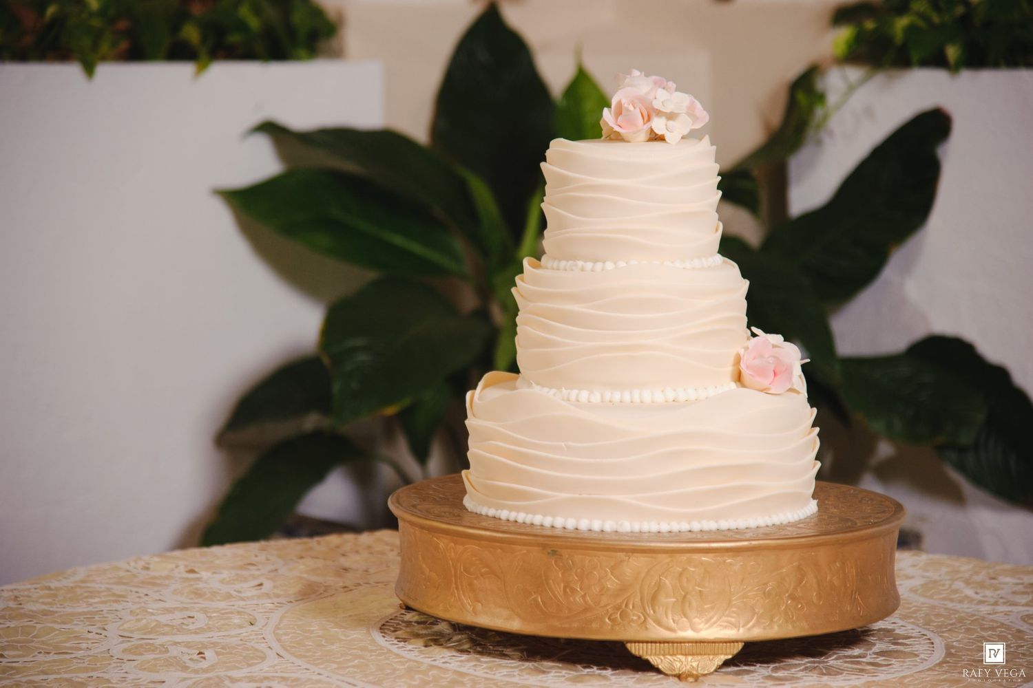 Elegant three-tier white wedding cake with ruffled frosting and pink roses on a gold cake stand.