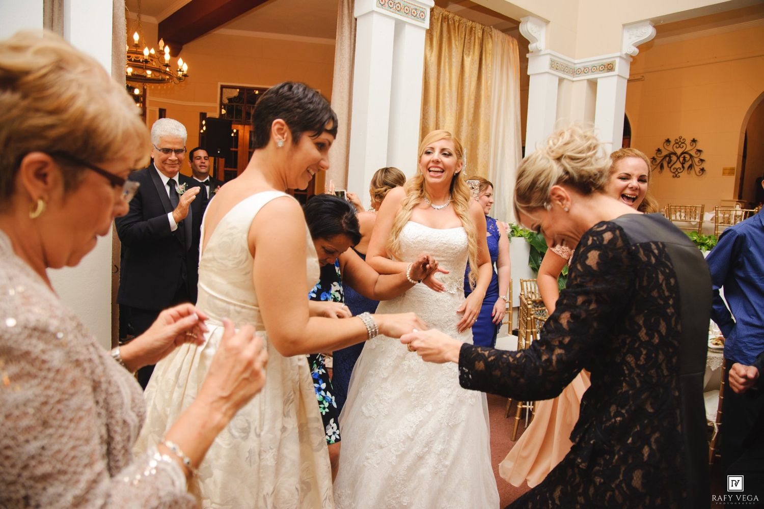 Two brides in white wedding gowns dance joyfully with guests at their wedding reception indoors.