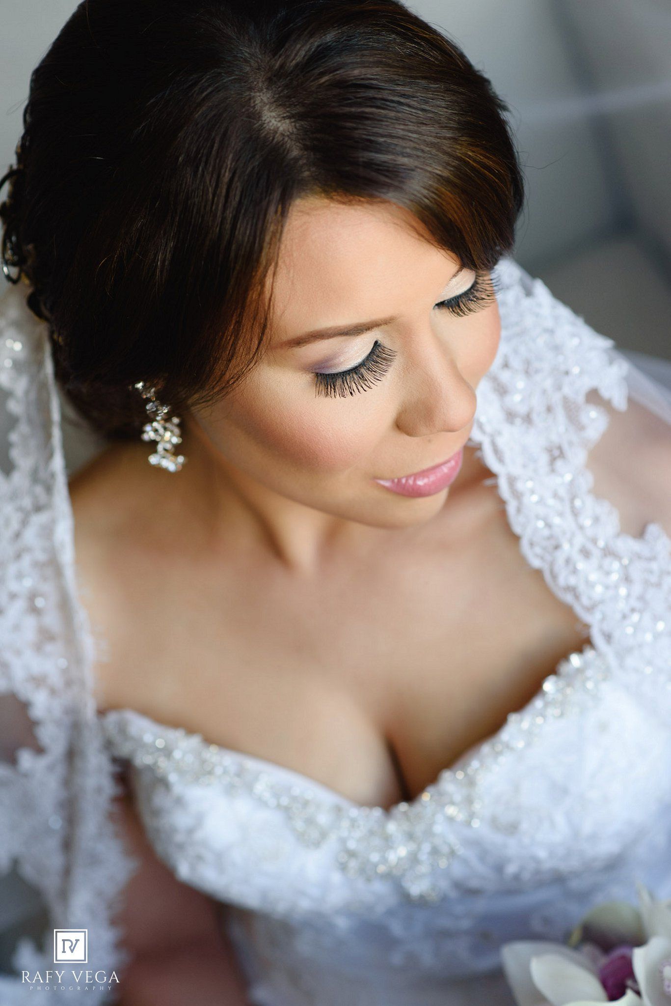 Elegant bride in lace wedding gown with pearl earrings and veil, wearing glamorous makeup with long lashes.