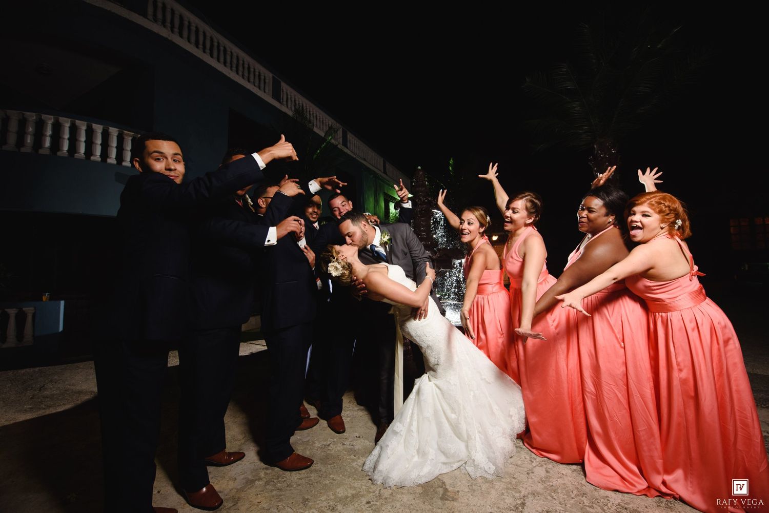 Wedding party celebrating outdoors at night, bridesmaids in coral gowns cheering as couple shares a dip kiss.